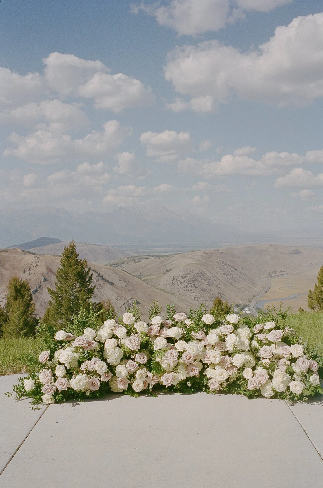 A floral arrangement of white and light pink roses and white hydrangeas on a table outdoors with a mountainous landscape, green trees, and a partly cloudy sky in the background.