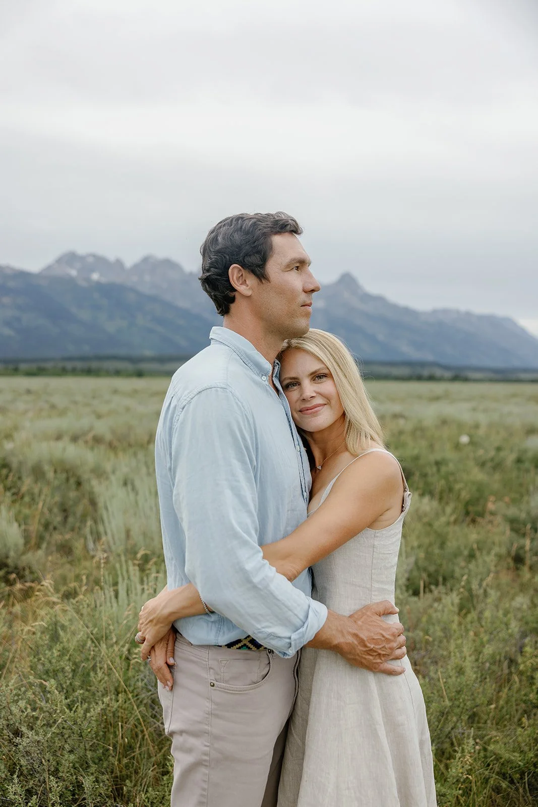 A man and woman embrace in a grassy field with mountains in the background. The woman rests her head on the man's chest, smiling softly; the man looks ahead with a serious expression.