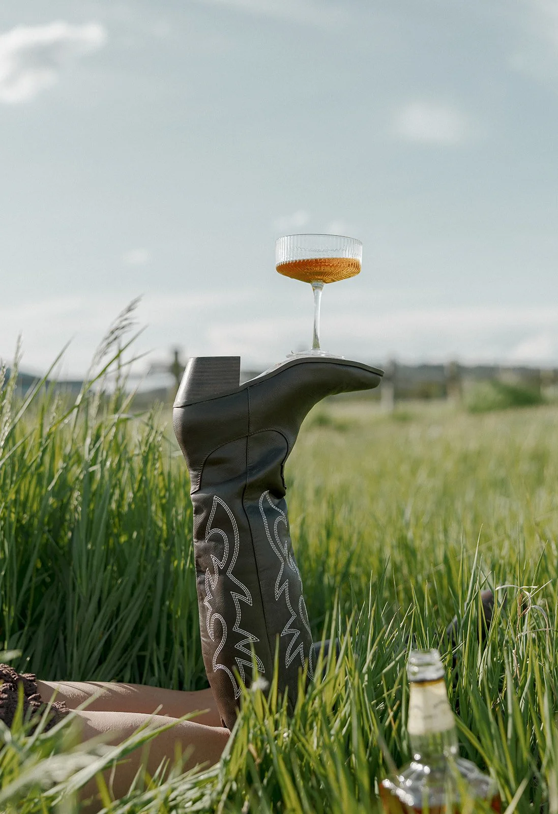 A grassy field with a golf club sock used as a stand, holding a glass of amber beverage. A skateboard is partially visible on the ground, and a clear bottle is in the grass nearby, under a partly cloudy sky.