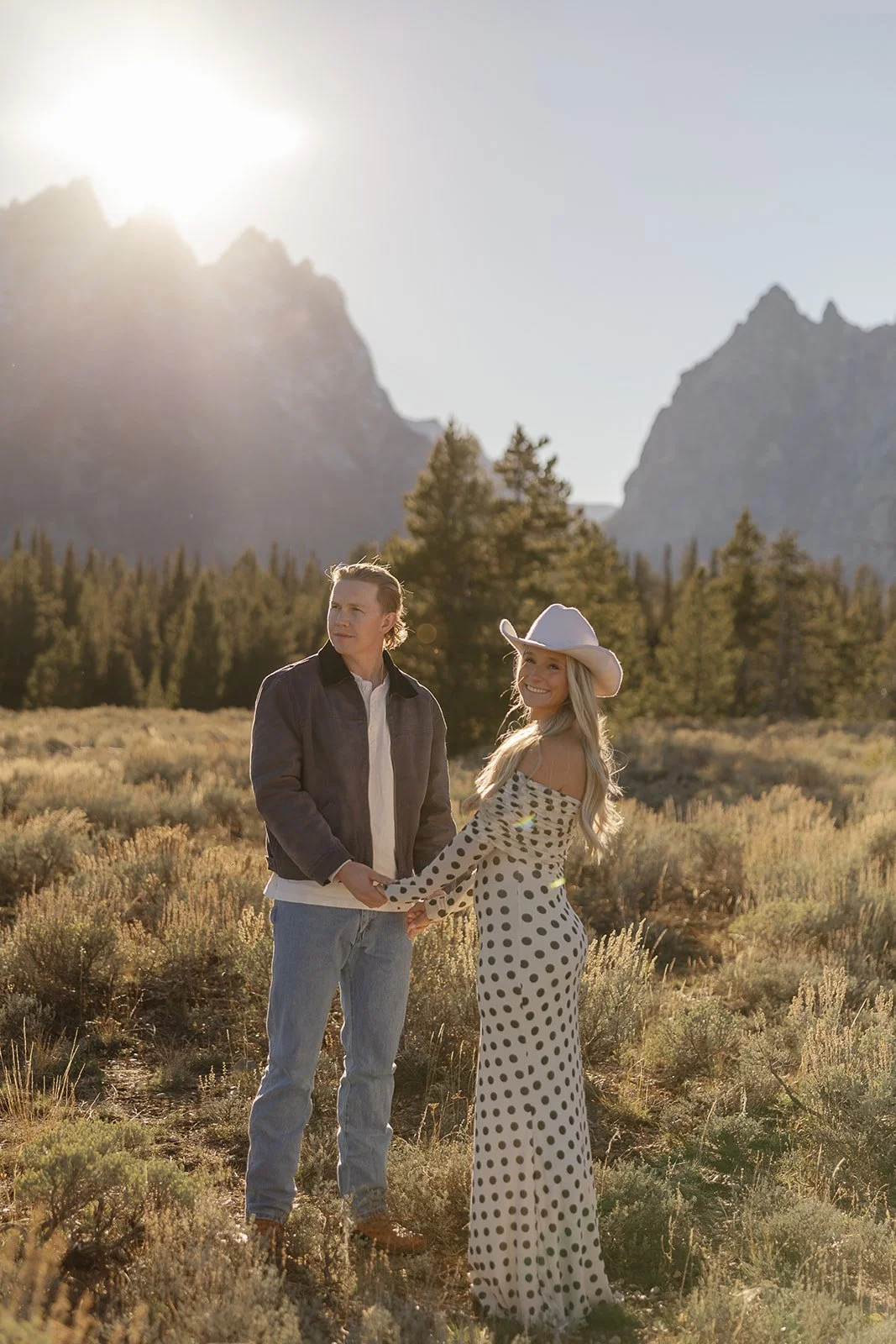 A couple holding hands in a field with mountains and trees in the background, sunlight shining from behind.