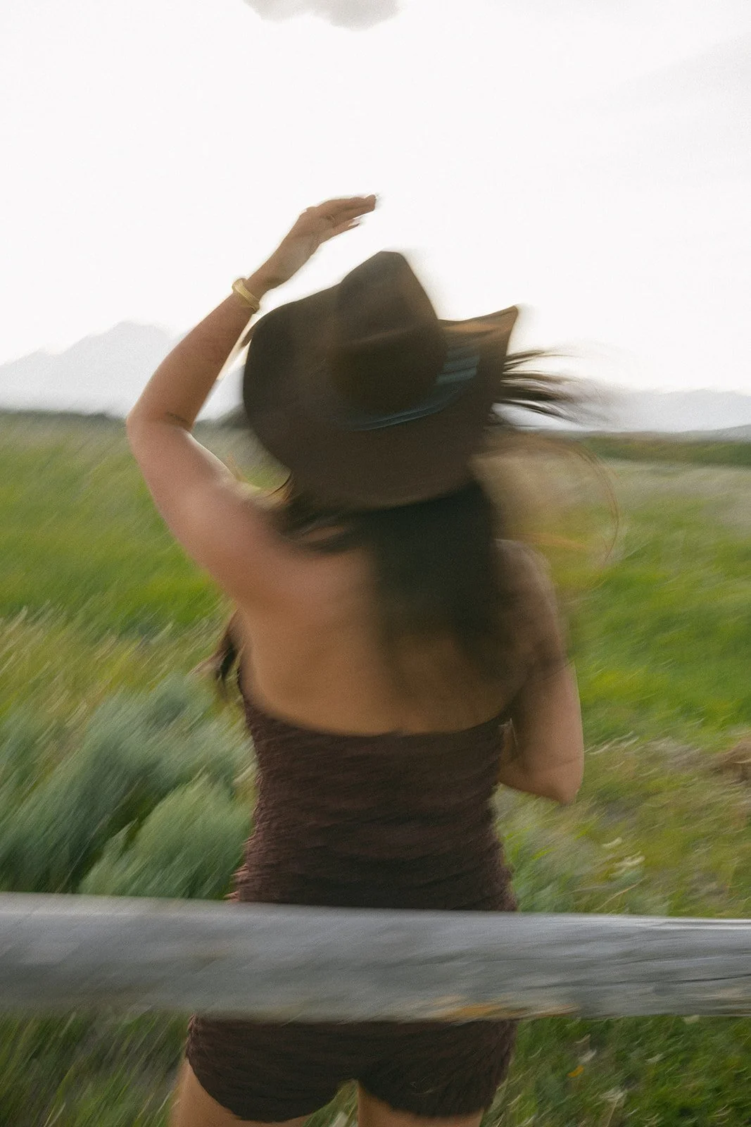 A woman in a black dress and hat standing outdoors with her back to the camera, resting her right hand on her head, with blurred green fields and mountains in the background.