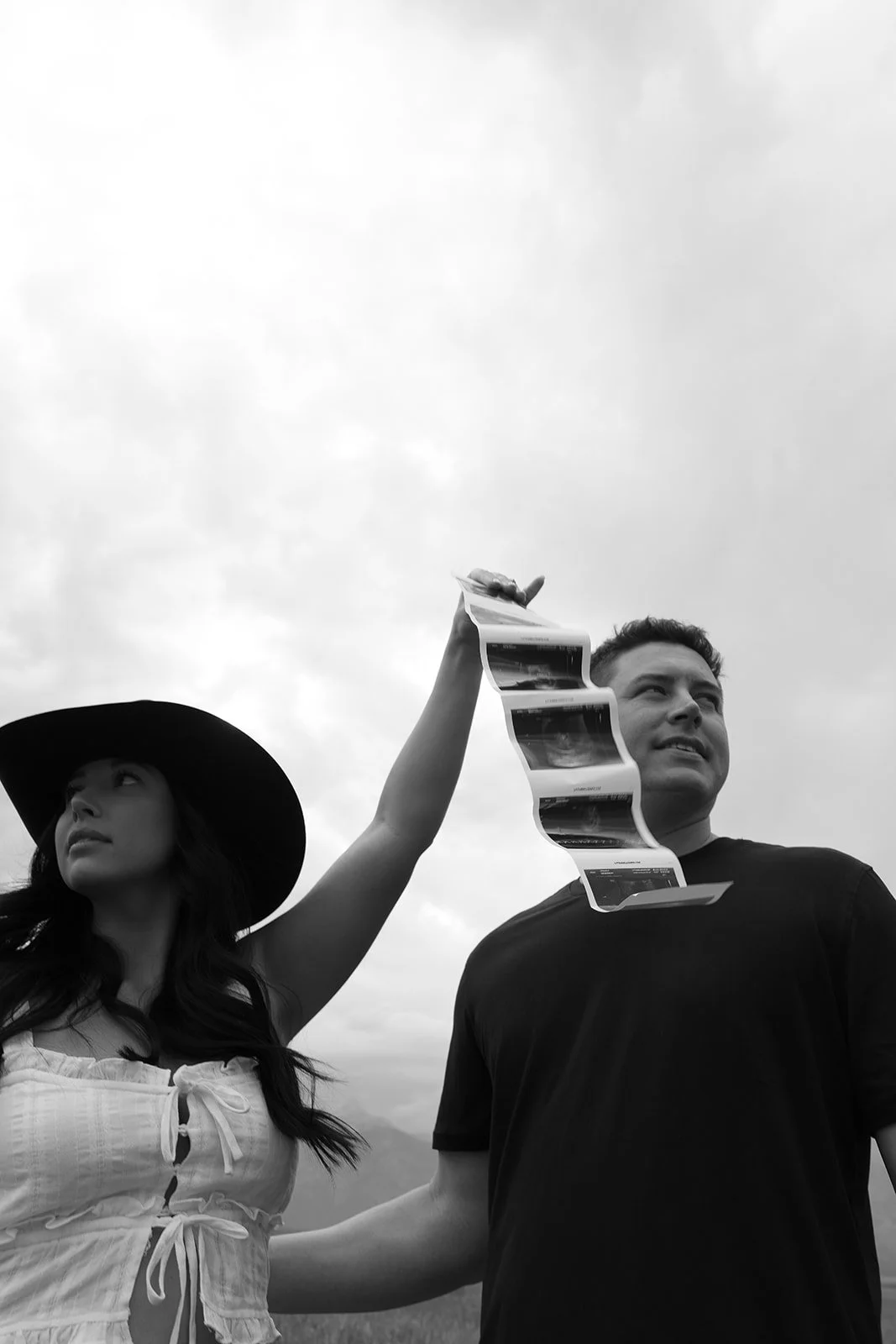 A woman wearing a large hat holding a chain of photographs or film strips over a man's shoulder outdoors.