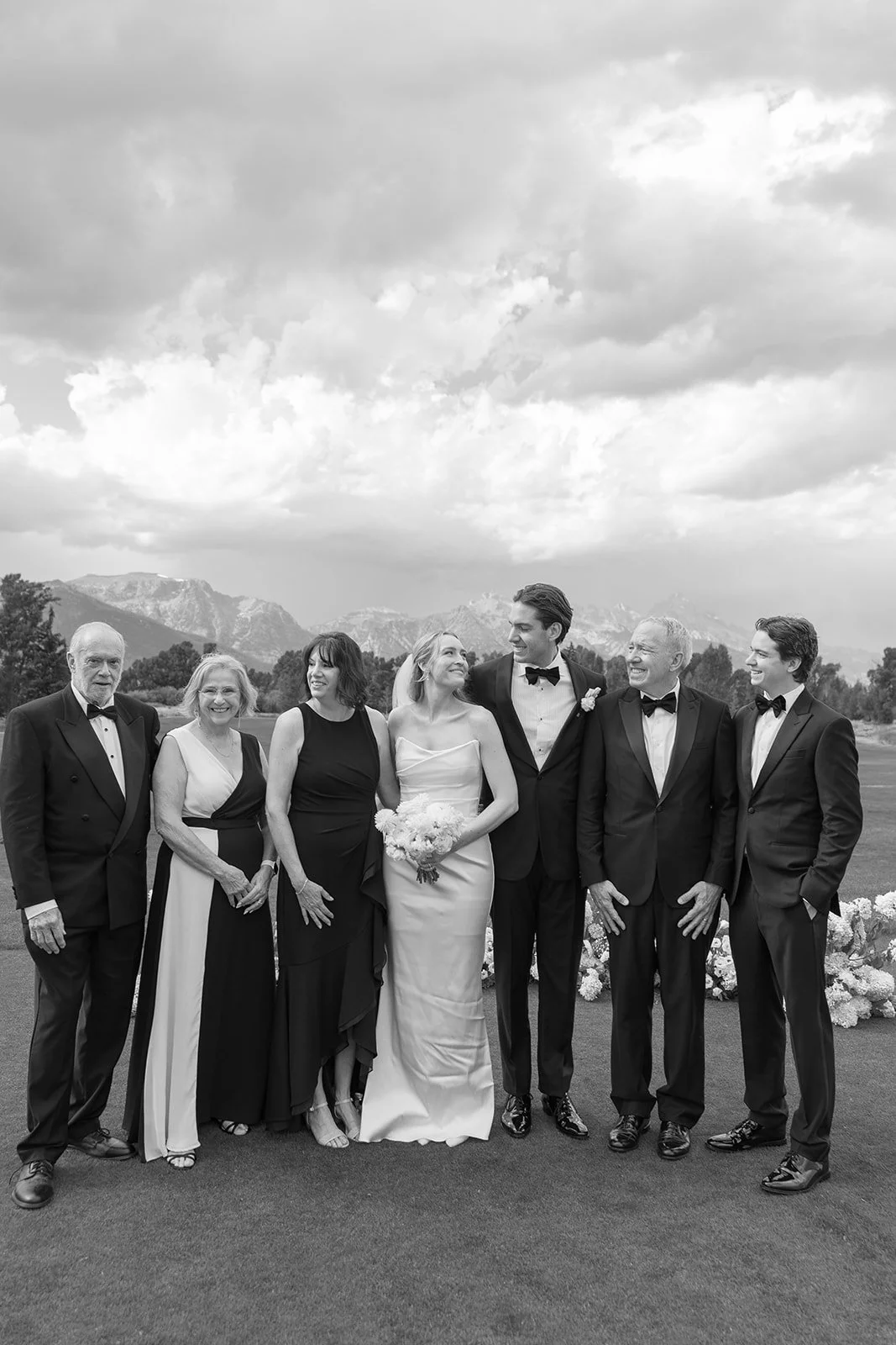 Black and white wedding photograph of a group of seven people, including a bride and groom in the center, standing outdoors with mountains and cloudy sky in the background, all dressed in formal attire.