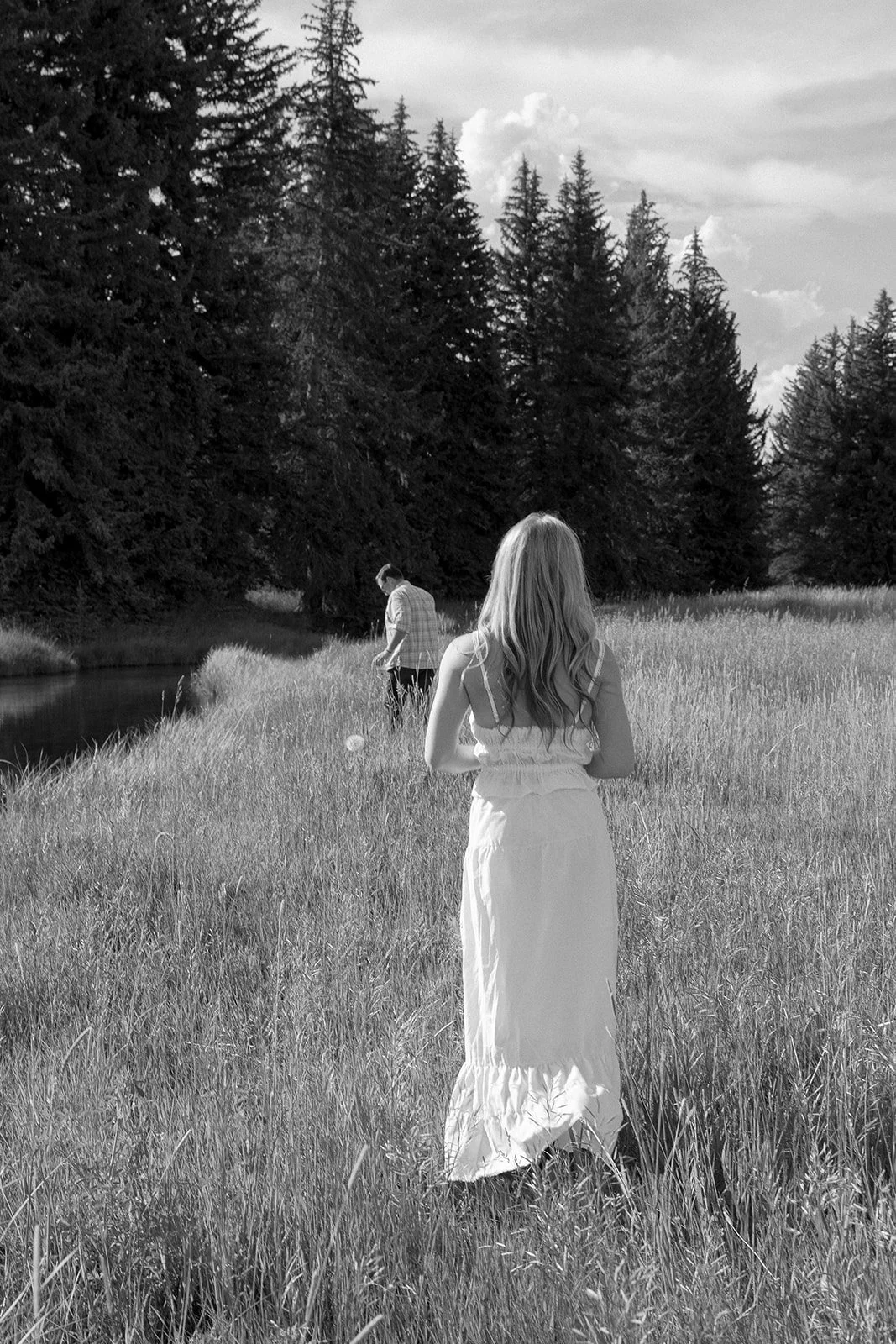 A black and white photograph of a girl in a long white dress walking through a grassy field next to a stream, with a man in a checkered shirt further ahead near the water, surrounded by tall trees and a cloudy sky.