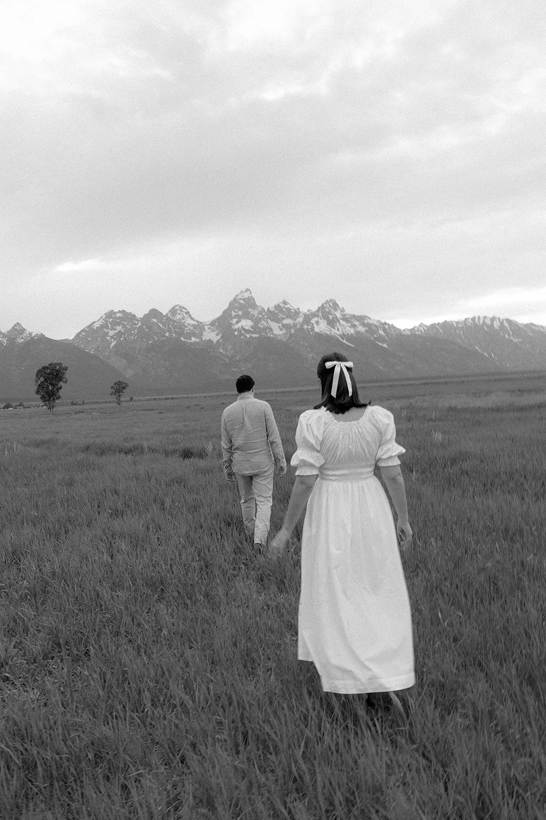 A black-and-white photo of a couple walking in a grassy field towards mountains in the distance, with one woman in a white dress and the man in a light-colored suit.
