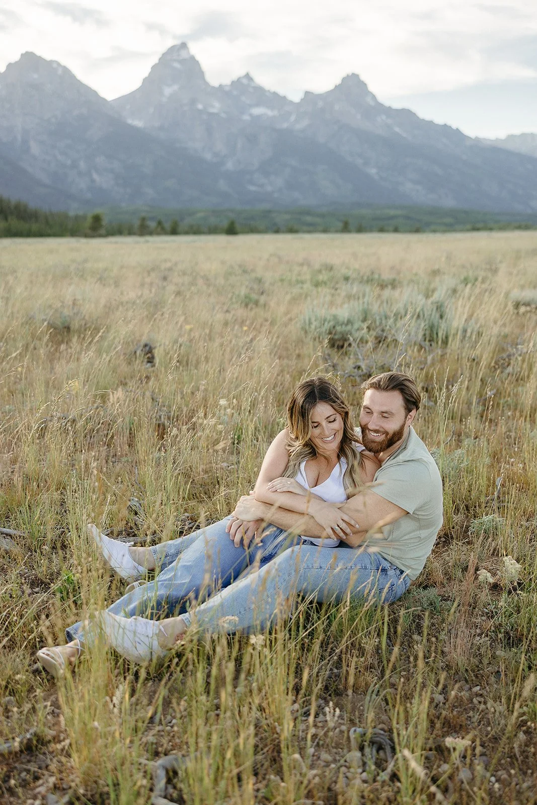 A happy couple sitting and cuddling in a grassy field with mountains in the background.