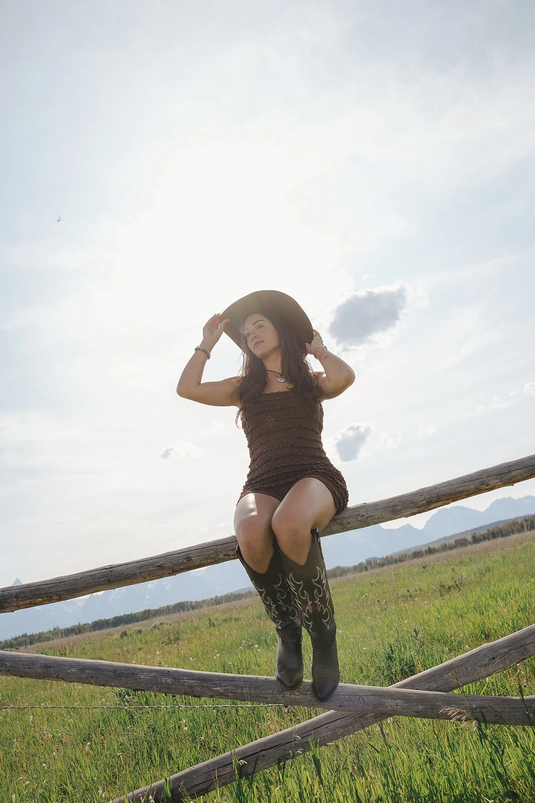 A woman sitting on a wooden fence in a grassy field, wearing a black hat, a brown dress, and tall black boots, with mountains in the background and the sun shining brightly.