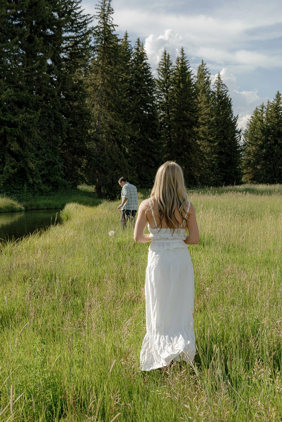 A woman with long blonde hair in a white dress walking through a grassy field near a small pond, with a man in the background by the water, surrounded by tall trees and a partly cloudy sky.