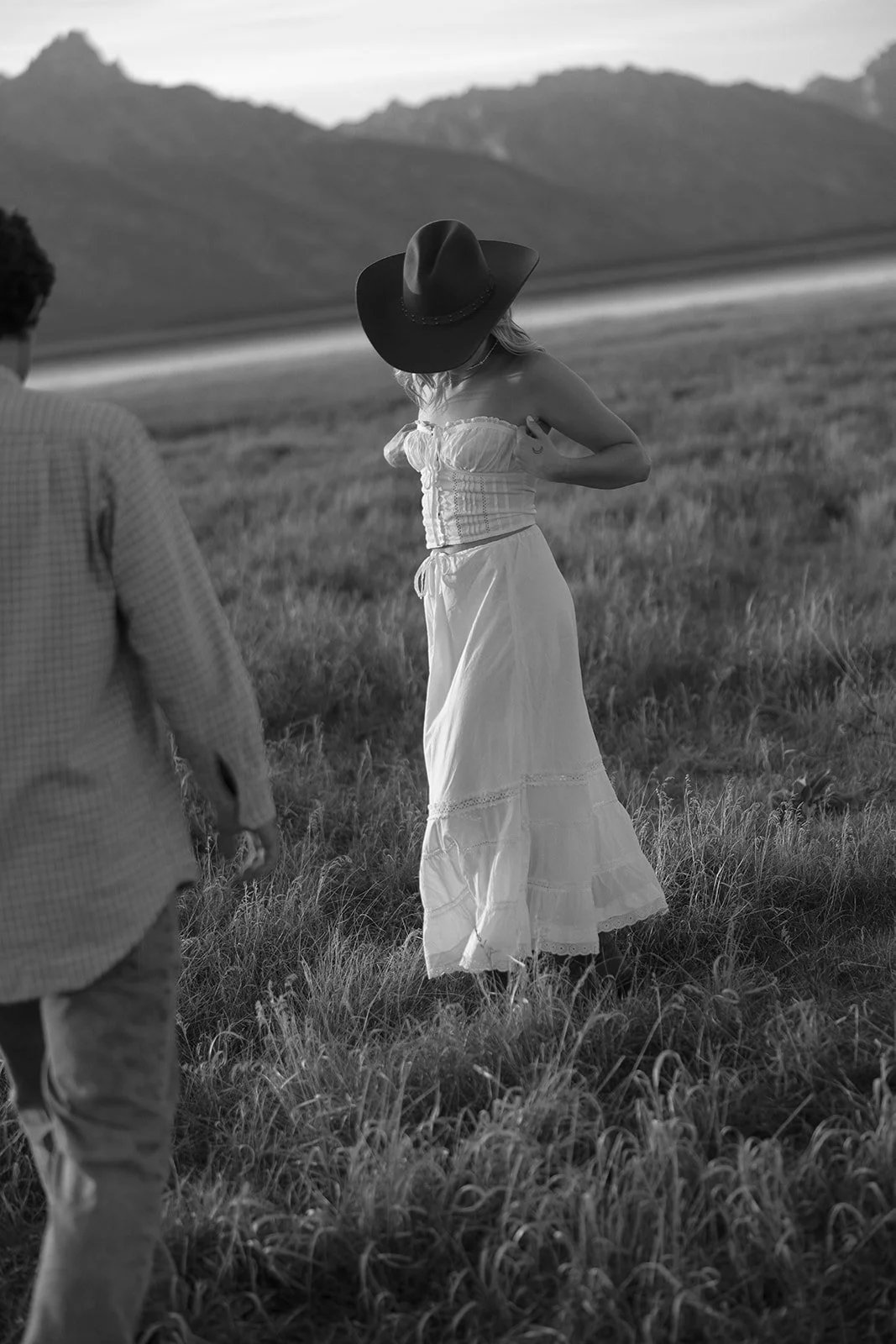 A woman in a white dress and large hat standing in a grassy field with mountains in the background, accompanied by a man.