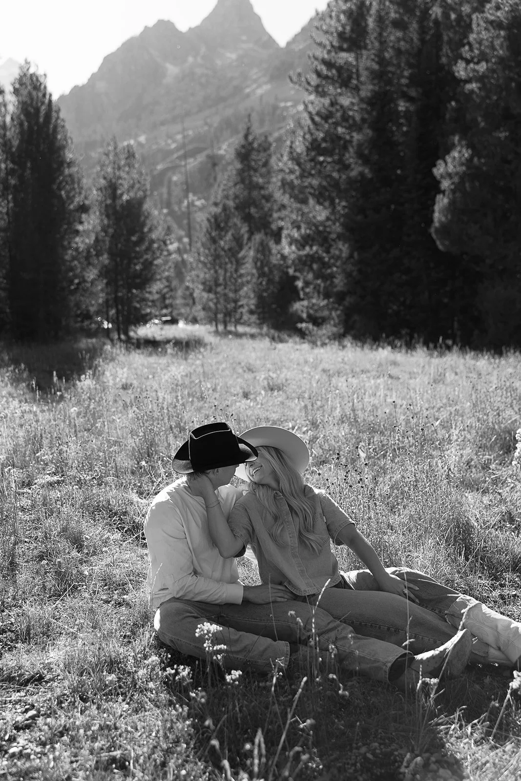 Two women sitting on a grassy field, wearing hats and smiling at each other, surrounded by trees with mountains in the background.