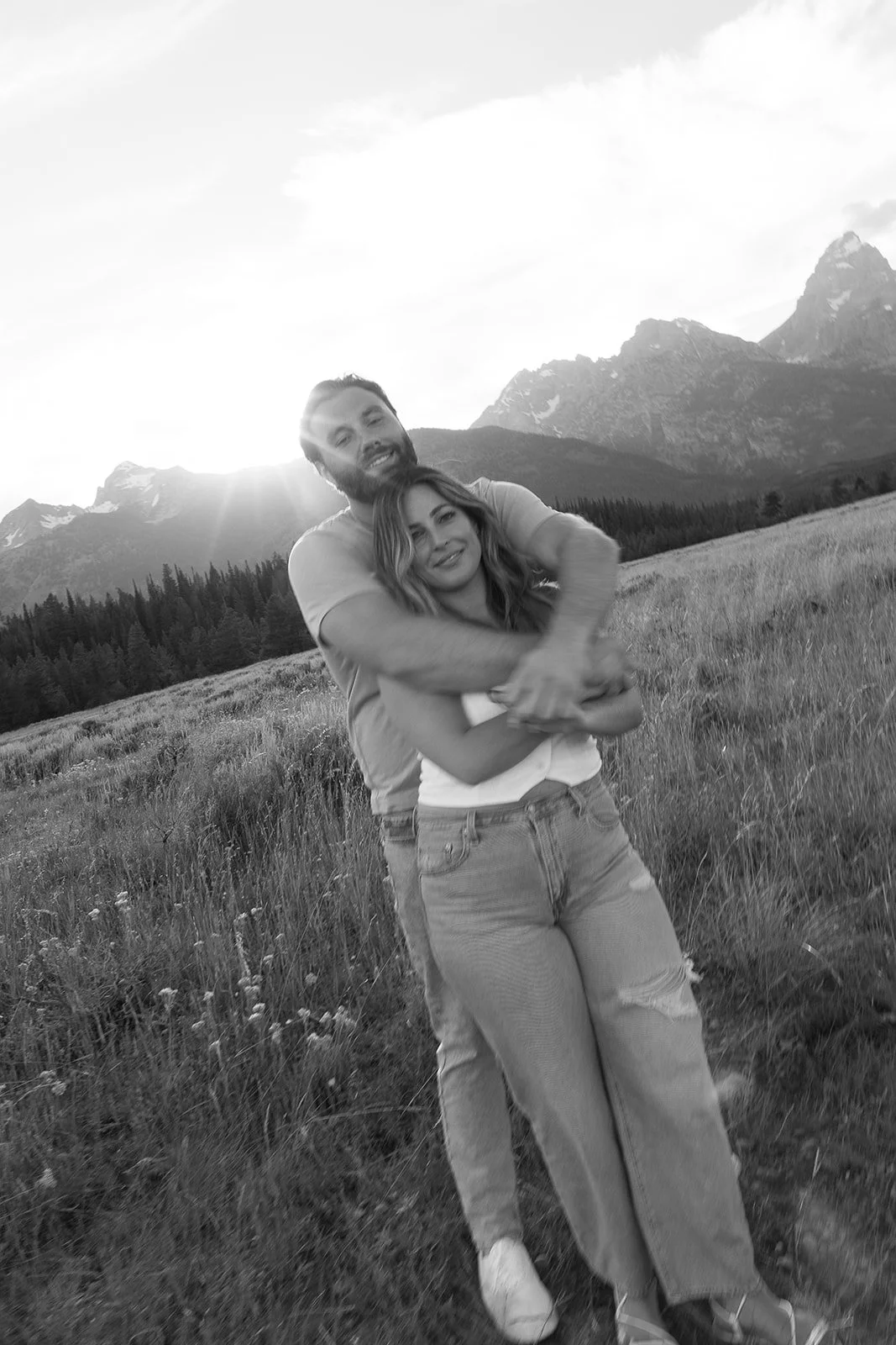 A black and white photo of a young couple embracing in an open field with mountains and a forest in the background, during sunset.