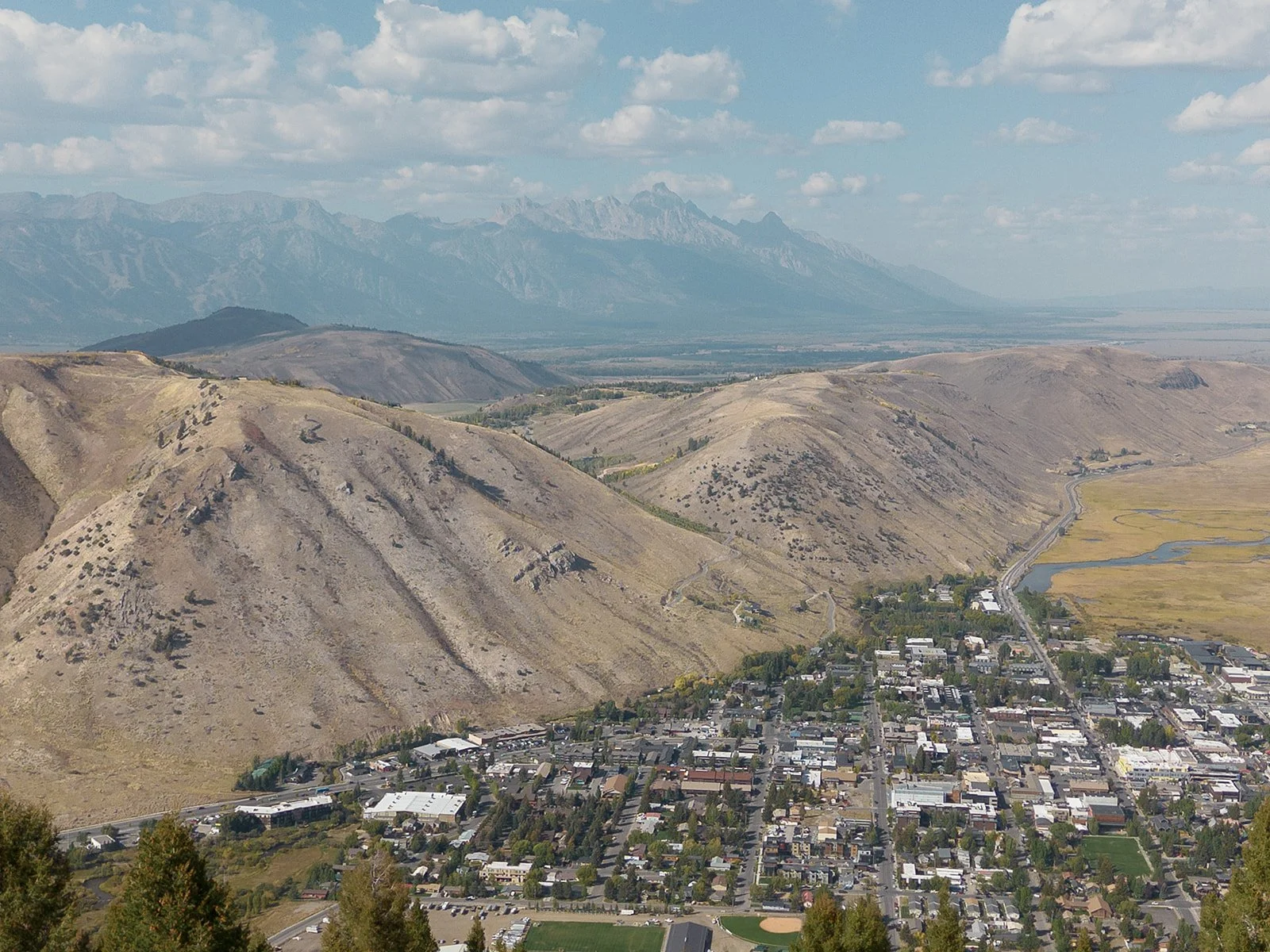 Aerial view of a small town nestled in a valley with rolling hills, mountain ranges in the background, and partly cloudy sky.