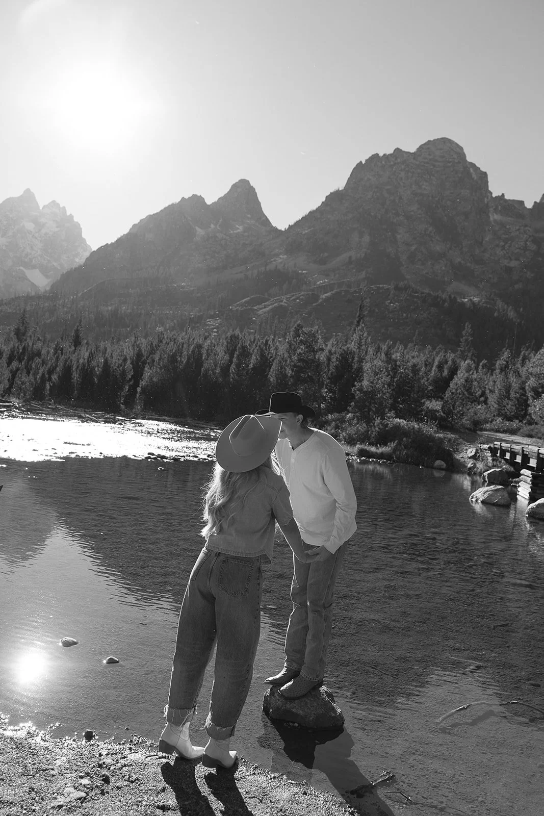 A couple holding hands while standing on a rock in a river, with mountainous landscape in the background, in black and white.