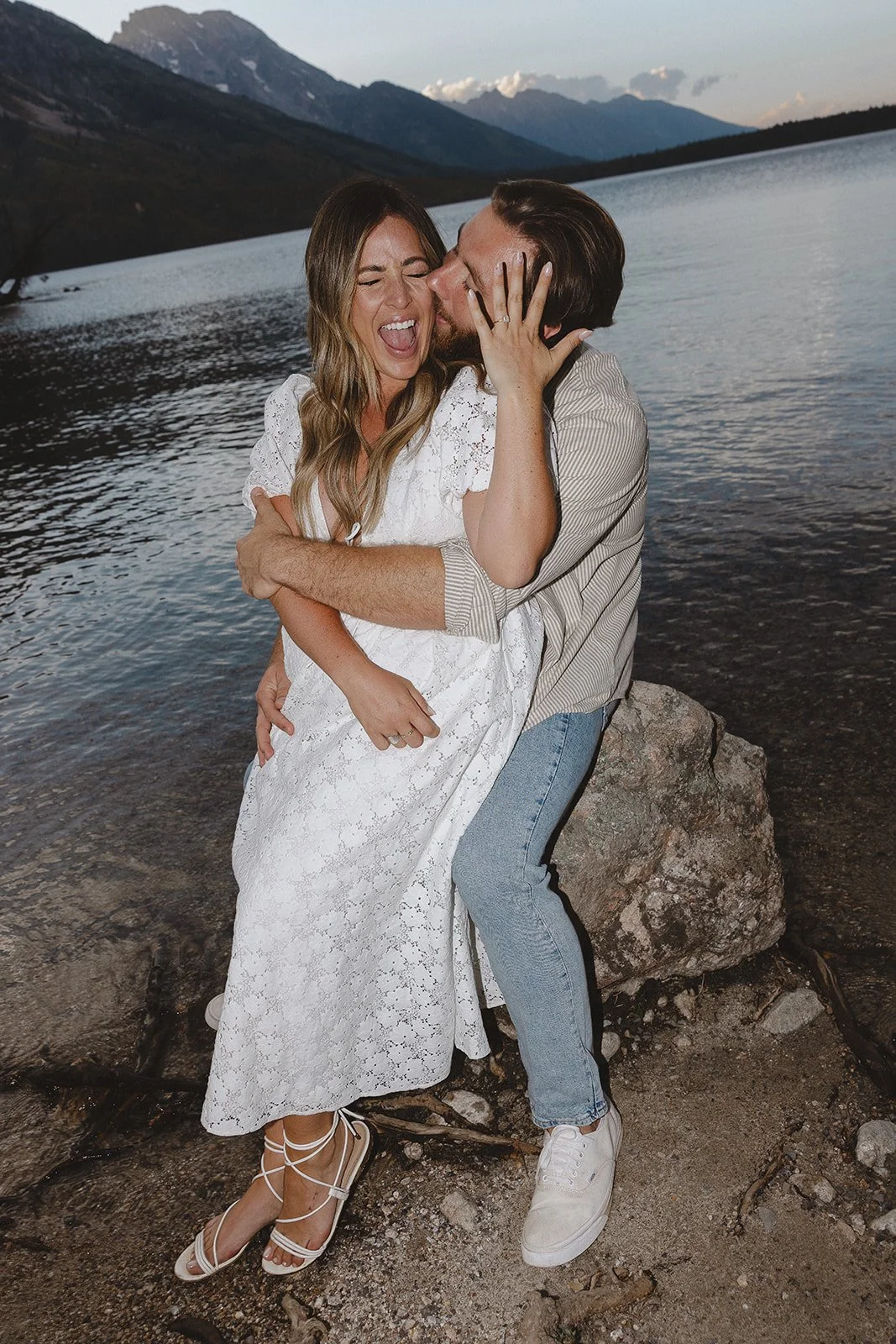 A couple embracing and laughing by a lakeshore with mountains in the background.