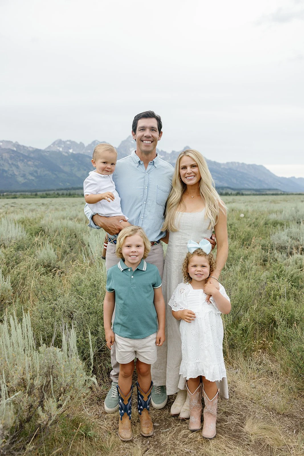 A family of five standing outdoors in a field with mountains in the background. The father holds a young boy, the mother stands next to him, with a young girl in a white dress and bow and another girl in a polo shirt and shorts in front.