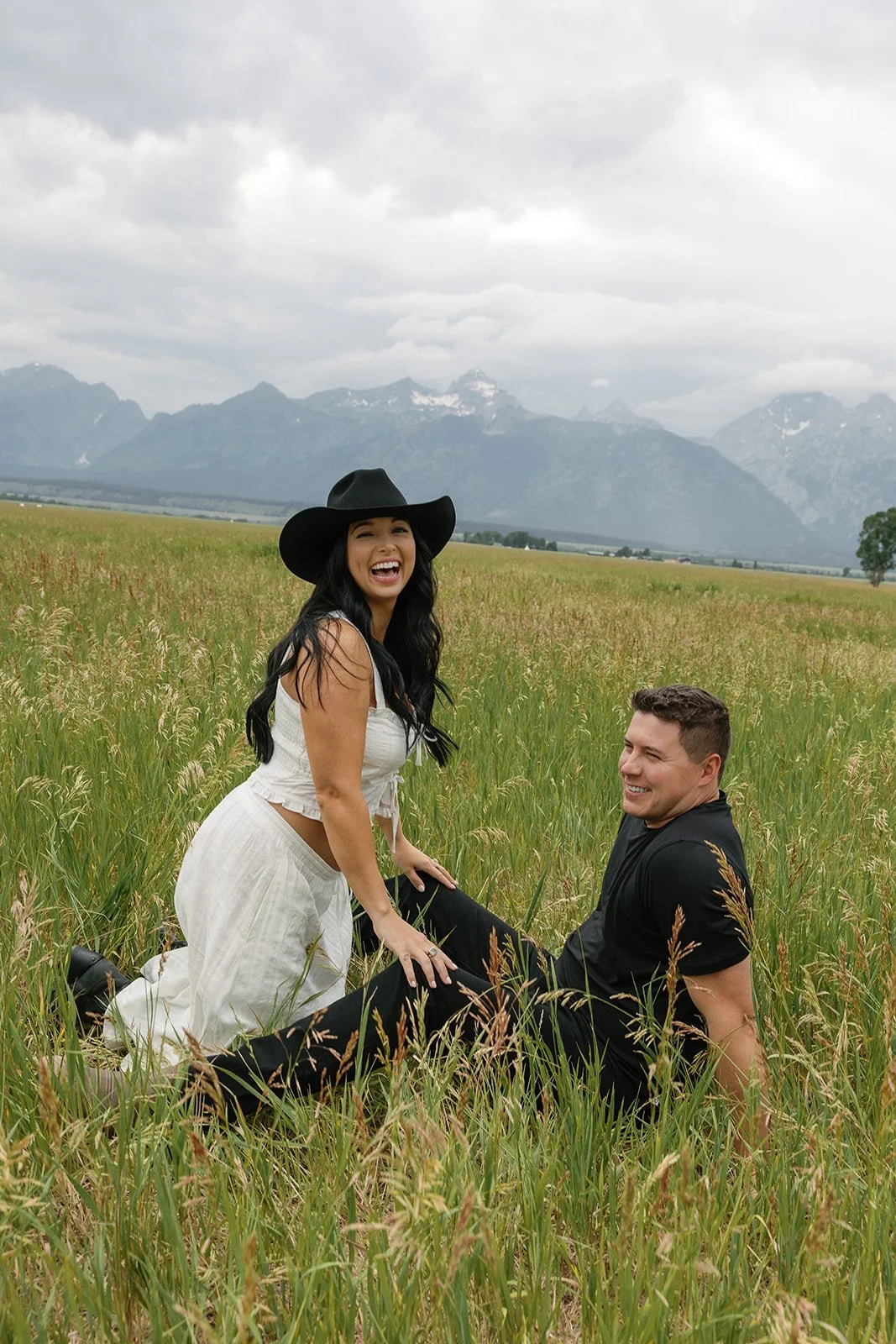 A woman wearing a black wide-brim hat and white dress, and a man in black shirt, sitting in a grassy field with mountains in the background, both smiling and enjoying the outdoors.