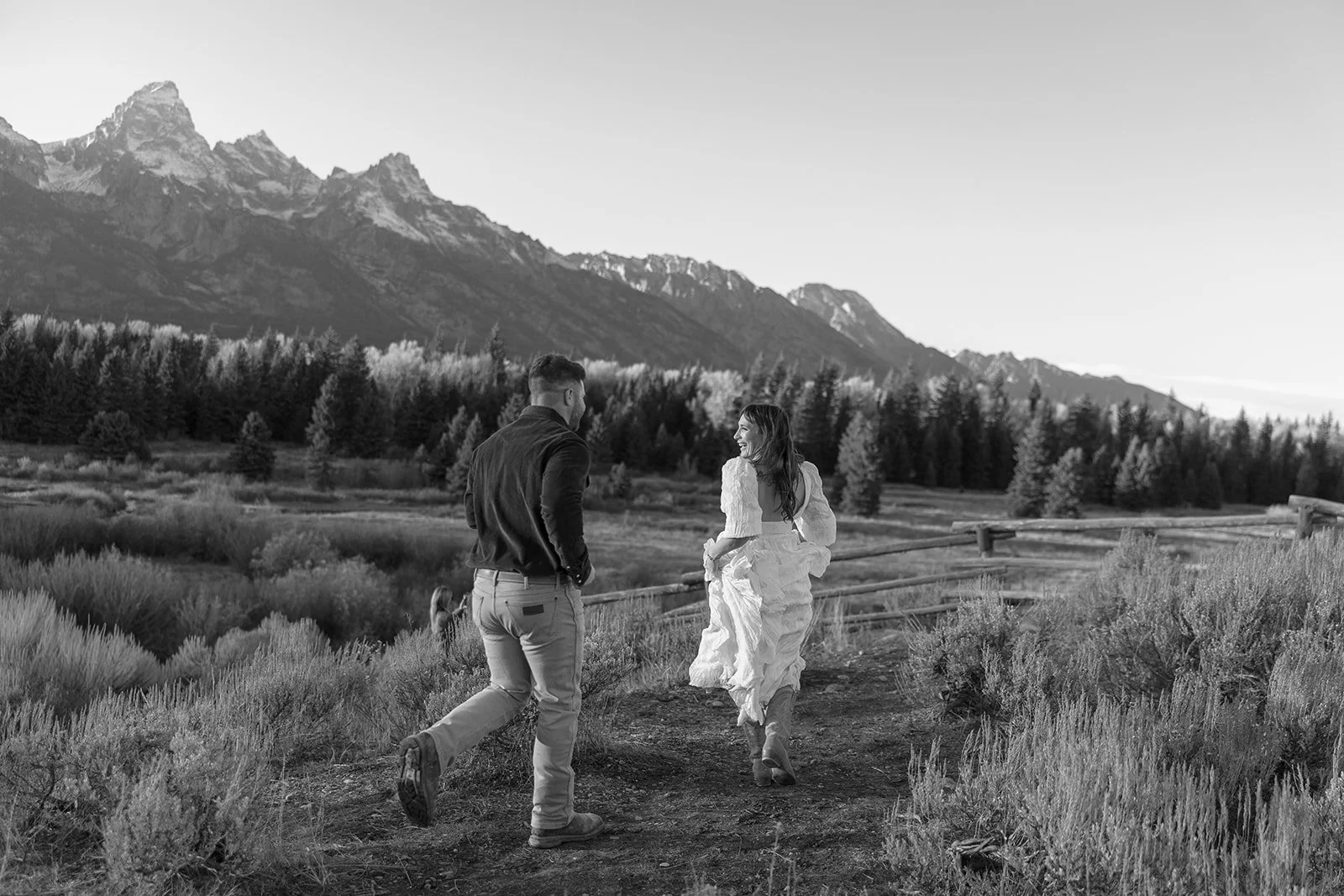A man and woman walking together on a dirt path in a landscape with mountains, trees, and shrubs, black and white photo.