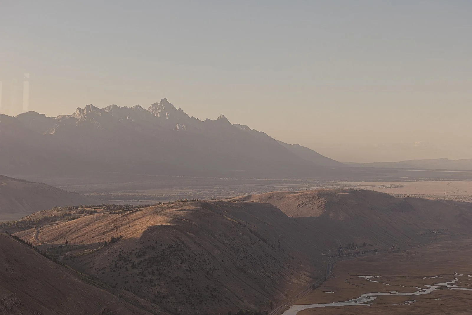 Mountain landscape with snow-capped peaks in the background, rolling hills and a winding river in the foreground during sunset.