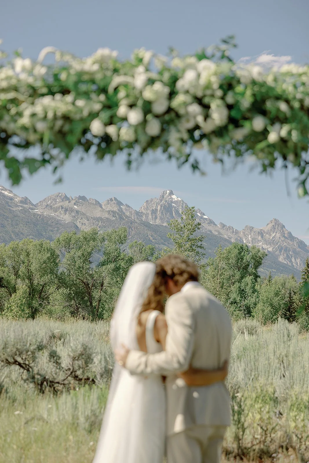 A couple dressed in wedding attire sharing an intimate moment in a scenic outdoor setting with mountains in the background, trees, and a cloudy sky.