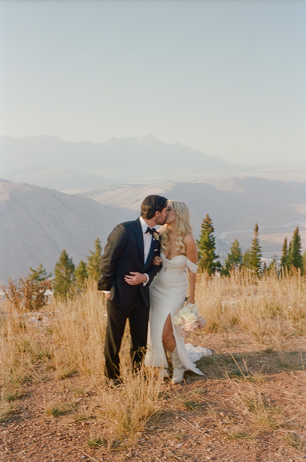 A bride and groom sharing a kiss in a scenic outdoor setting with mountains and trees in the background.