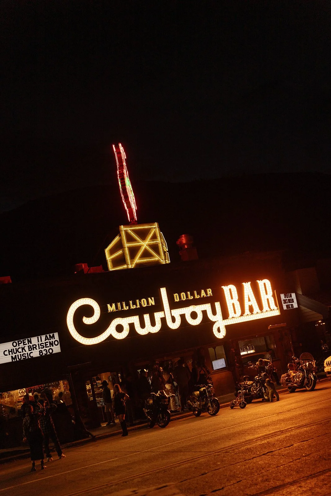 Nighttime image of the Million Dollar Cowboy Bar with people and motorcycles outside, neon sign illuminated, and fireworks in the sky.