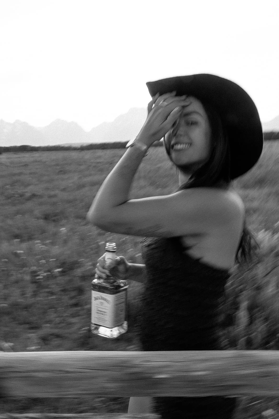 A woman wearing a wide-brimmed hat, holding a bottle of alcohol, smiling, and touching her forehead with her hand, outdoors in a rural area with mountains in the background.