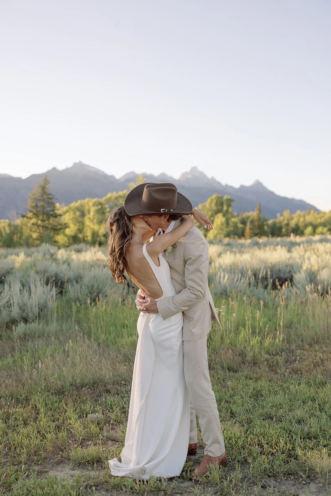 A couple in wedding attire sharing a kiss and embrace outdoors, with mountains and greenery in the background.