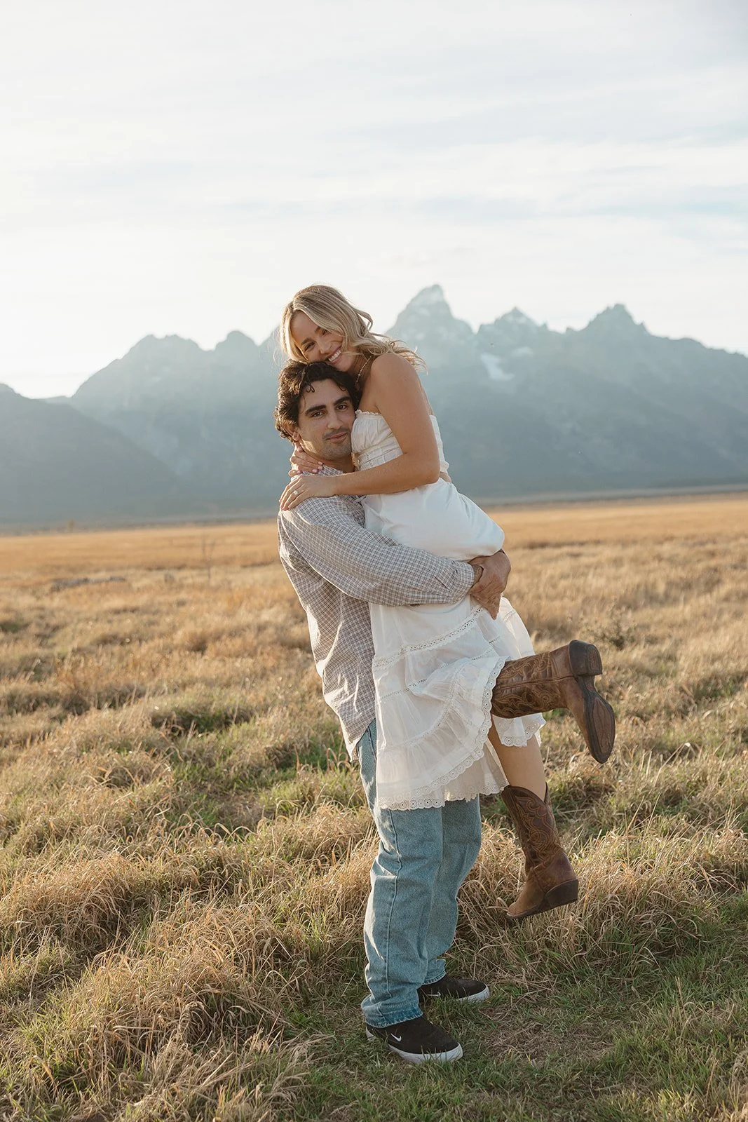 A couple in a grassy field with mountains in the background, the man lifts the woman, both smiling and dressed in casual Western-style clothing.