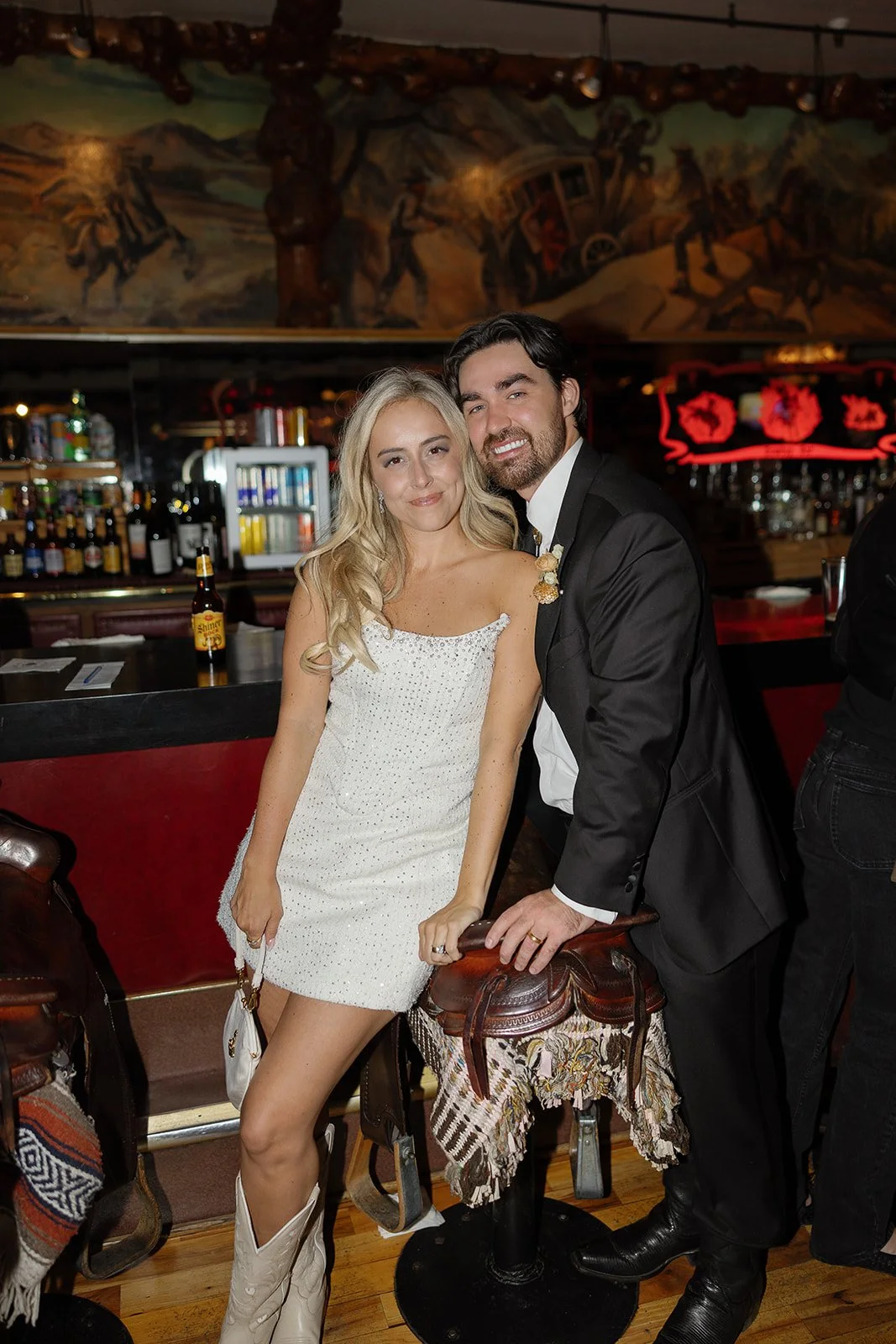 A woman in a white dress and cowboy boots standing next to a man in a black suit at a bar with a colorful mural painting in the background.