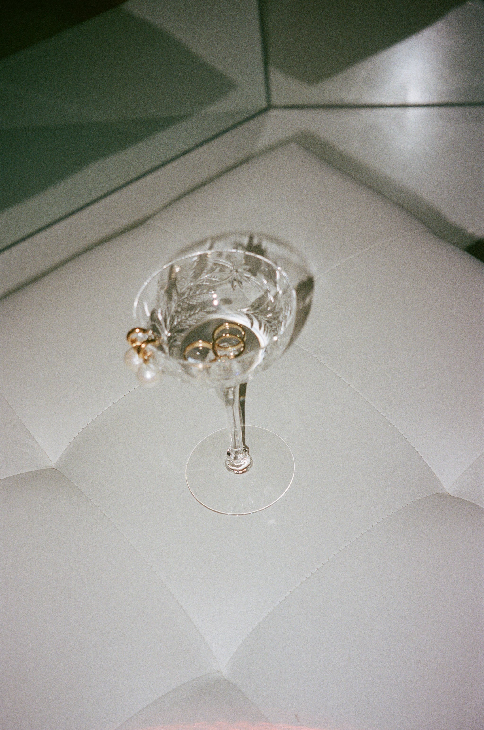Crystal champagne glass with gold rings and pearl earrings, on a white quilted surface.