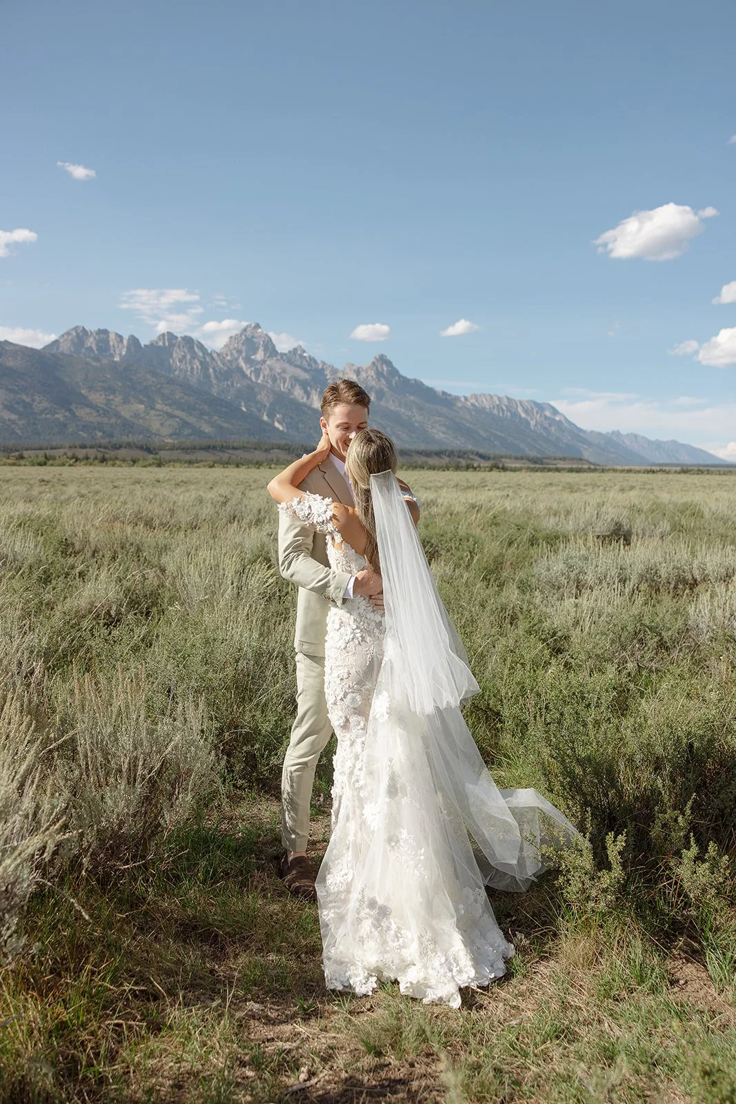 A bride and groom embracing outdoors in a grassy field with mountains in the background, under a blue sky with scattered clouds.