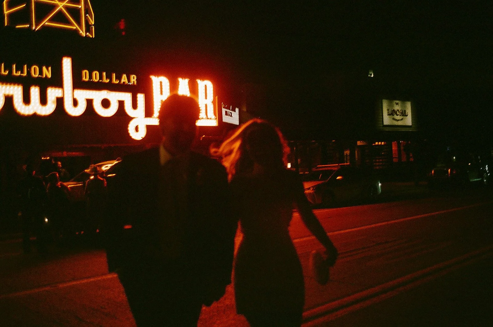 Silhouettes of a man and woman holding hands on a city street at night, illuminated by neon signs and streetlights.