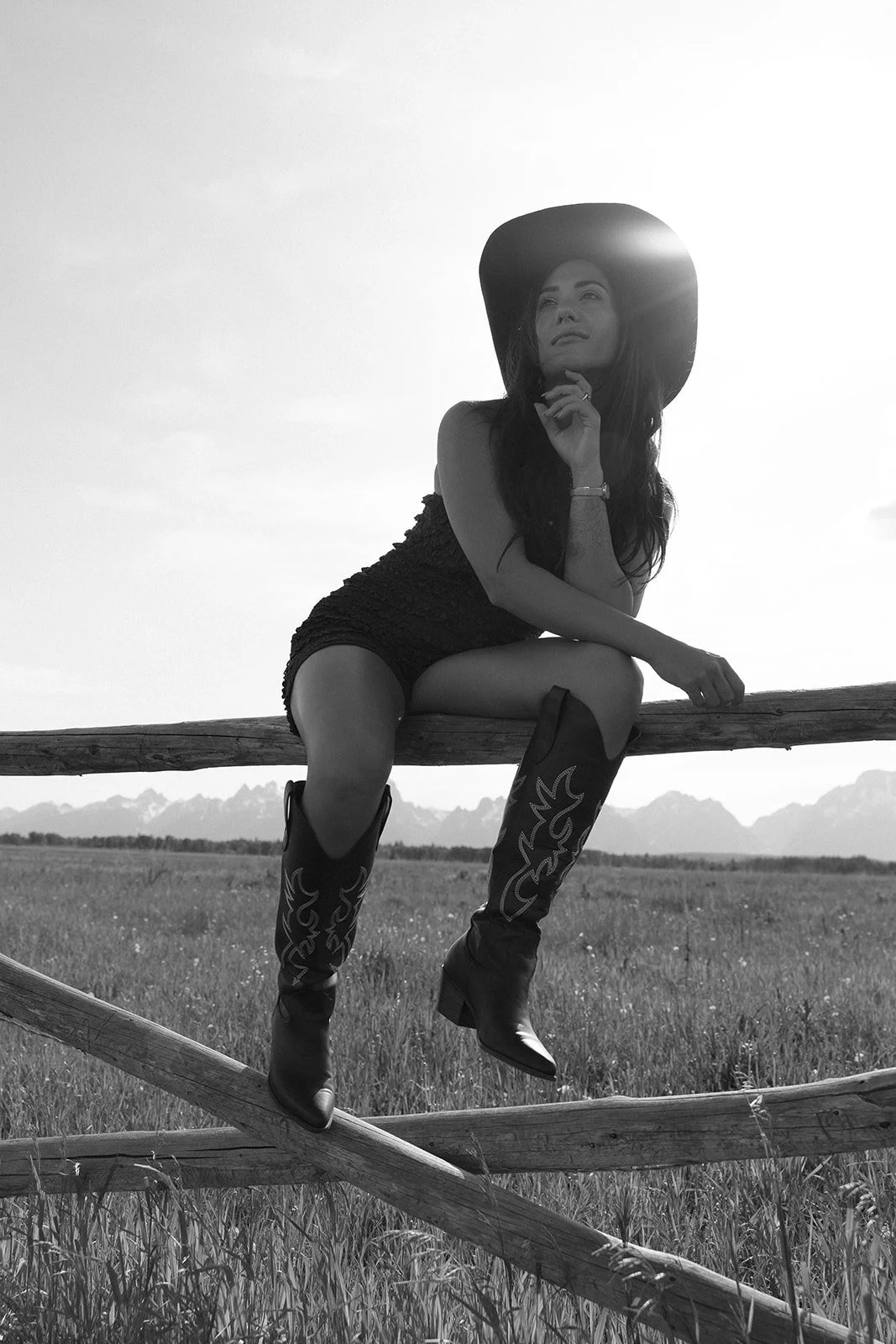 A woman sitting on a wooden fence in an open field, wearing a wide-brim hat, lace top, cowboy boots, looking into the distance, with a mountain range in the background, in black and white.