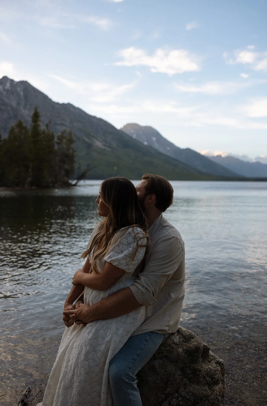 A couple hugging by a lake with mountains in the background during sunset or dusk.