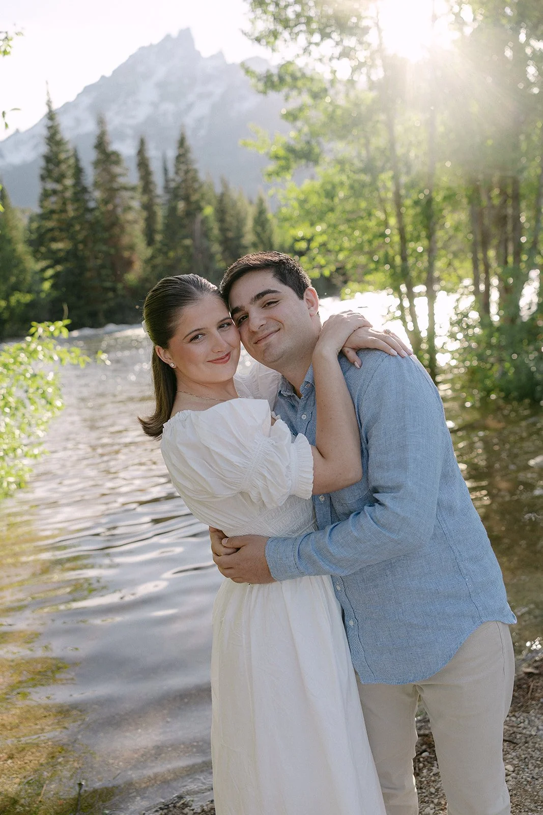 A smiling couple embracing near a river with a forest and mountain in the background, sunlight shining through trees.