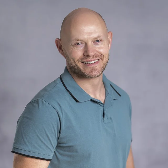 Ein lachender Mann mit einem blonden, kahl rasierten Kopf trägt ein blaues Polo-Shirt vor einem grauen Hintergrund.