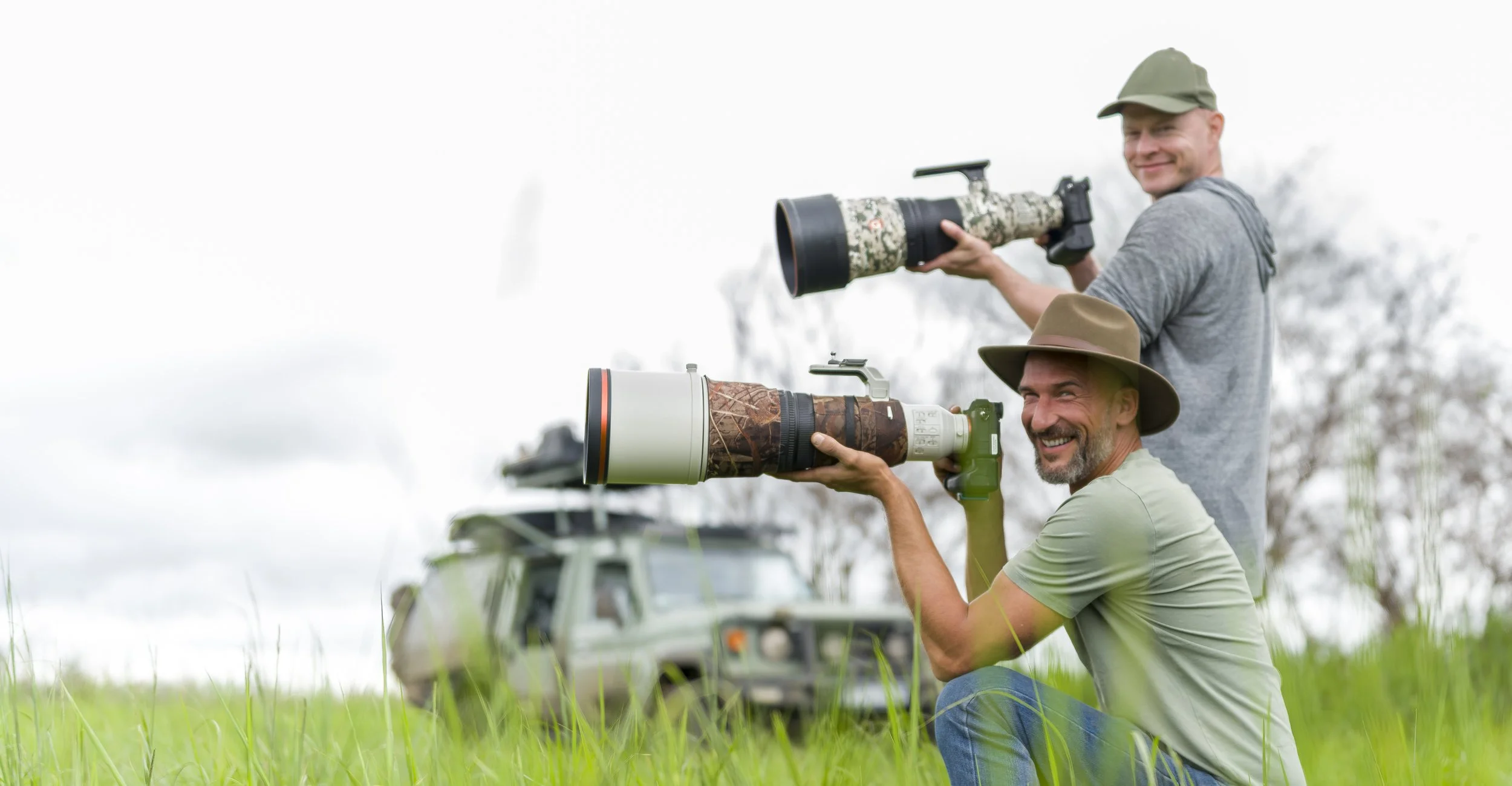 Zwei Männer mit Kameras mit Teleobjektiven im Freien, im Hintergrund ein geländegängiges Fahrzeug, beide lachen und tragen Hüte, die Szene spielt auf einer Wiese bei bewölktem Himmel.