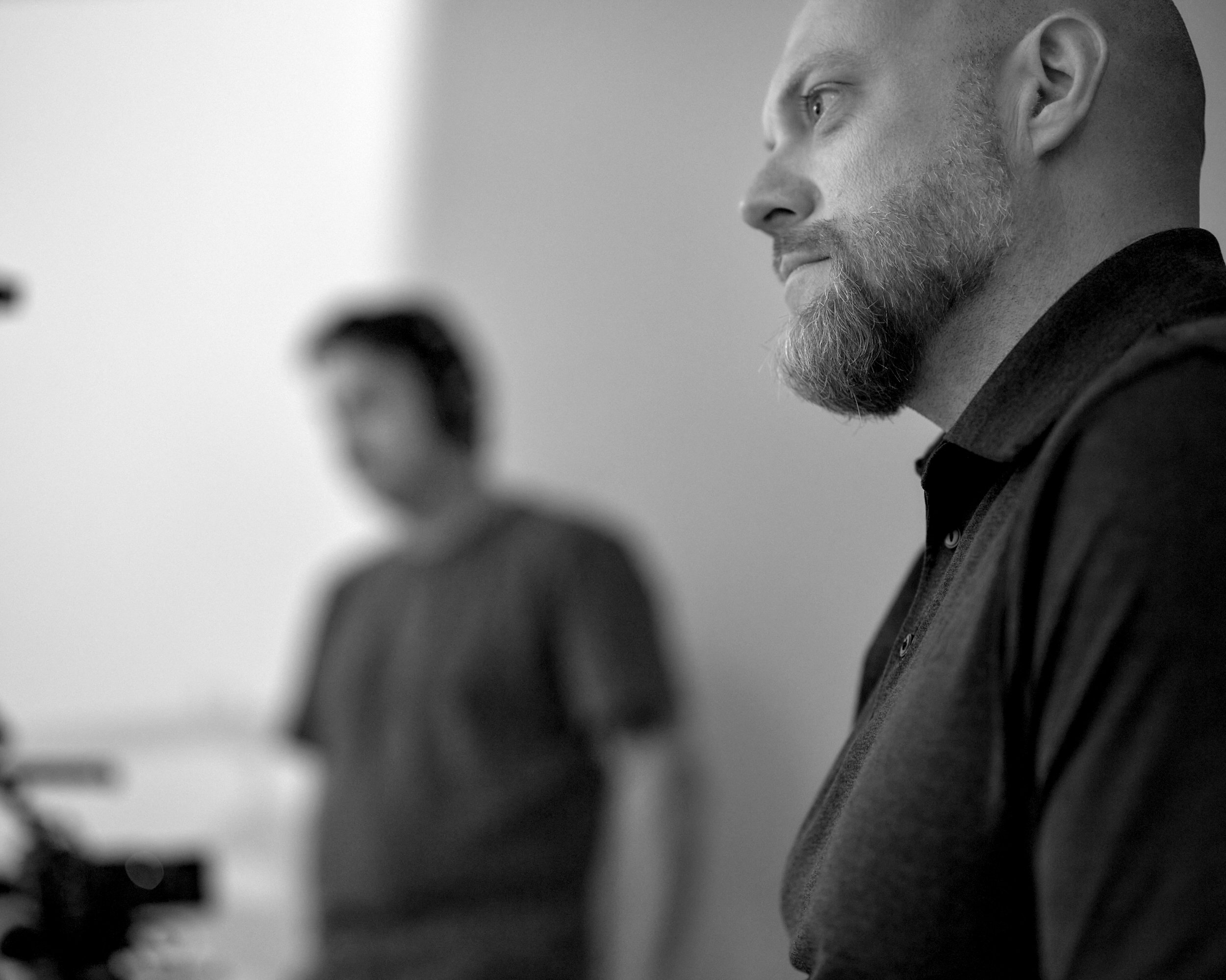 A close-up side profile of a man with a beard, in focus, with another person blurred in the background, both appearing to be in an indoor setting.