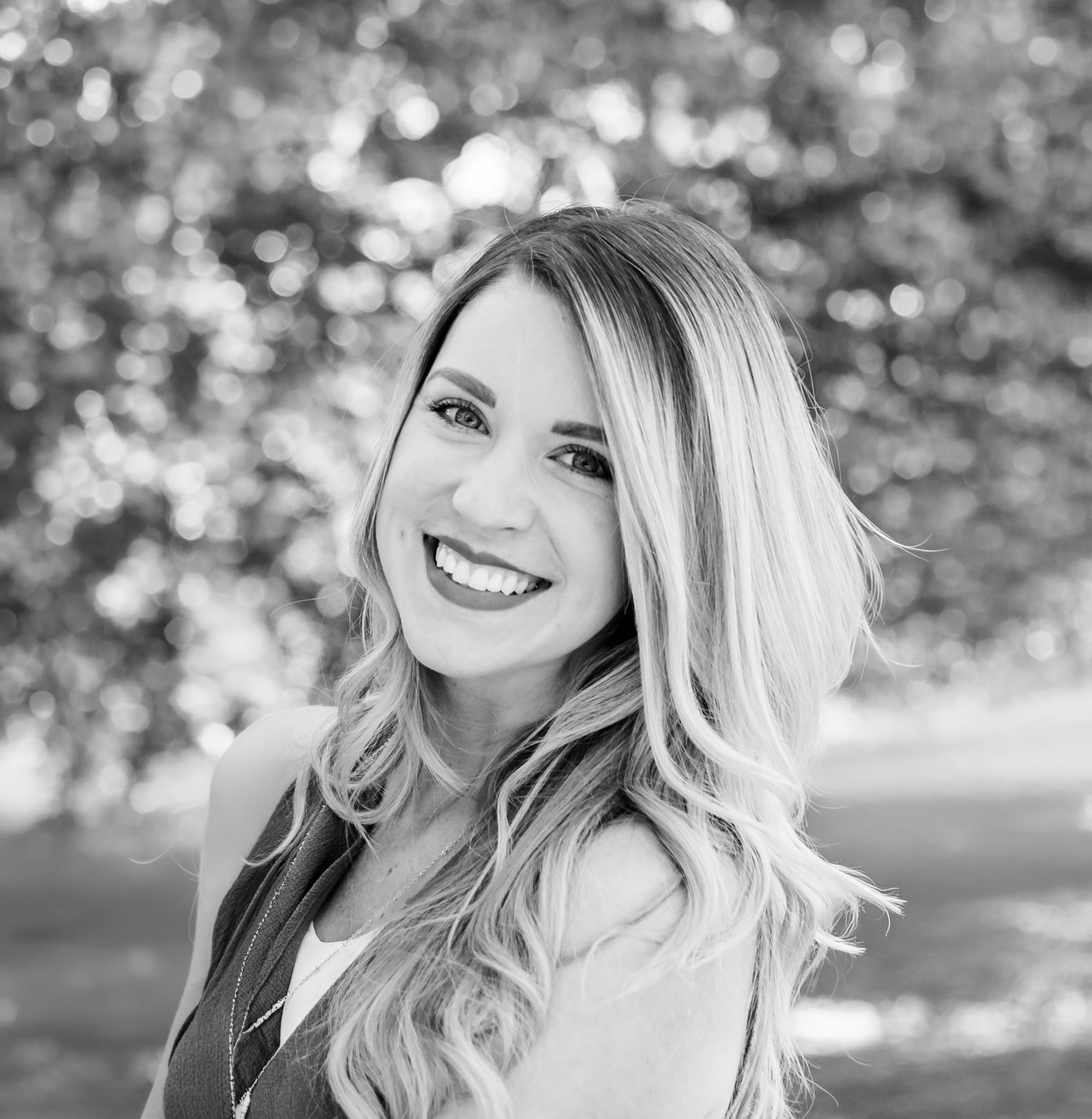 A smiling woman with long wavy hair standing outdoors with blurred trees in the background, photographed in black and white.