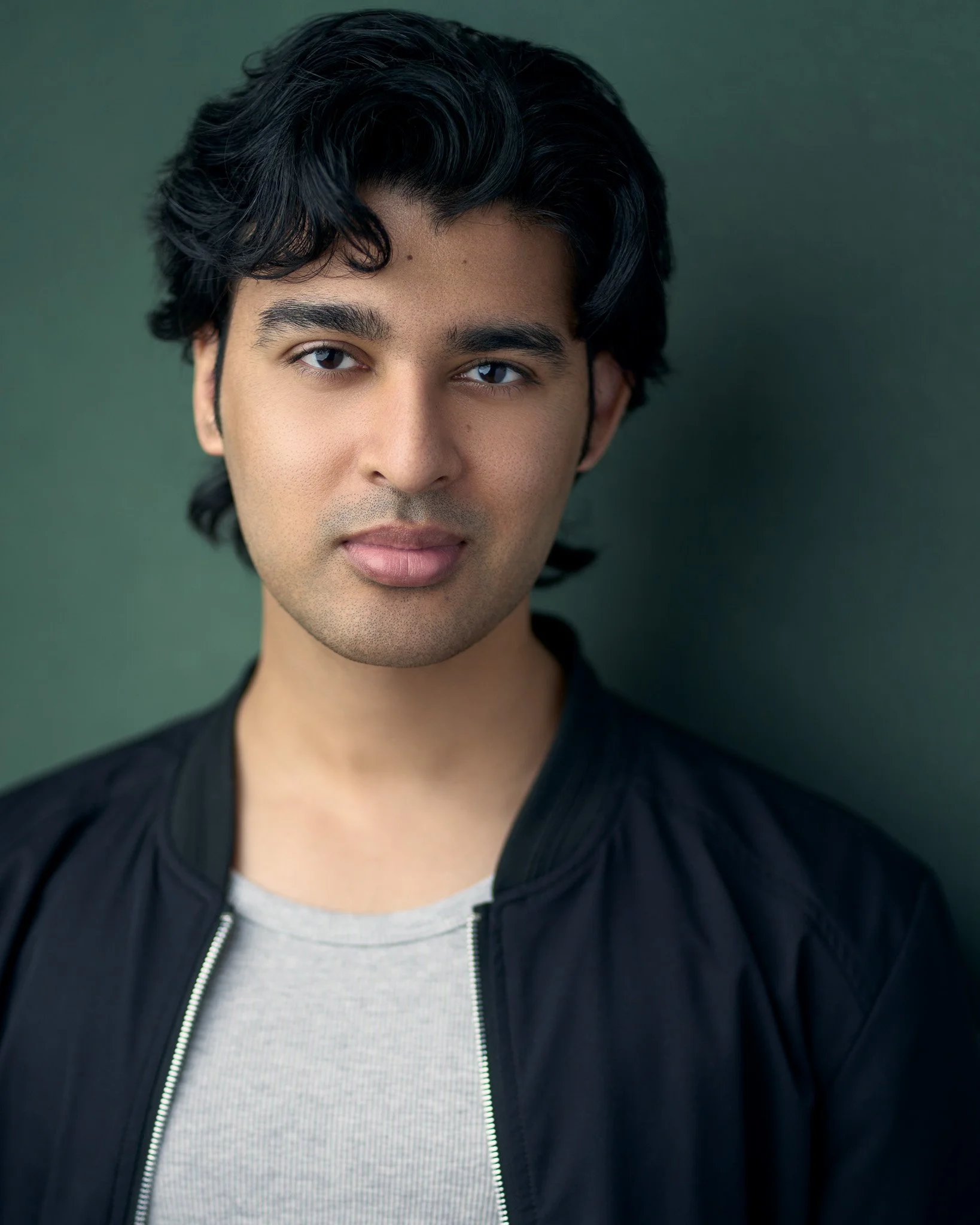 Acting headshot of a young man with wavy black hair and calm expression, wearing a gray shirt and black jacket, poses against a dark green textured background.