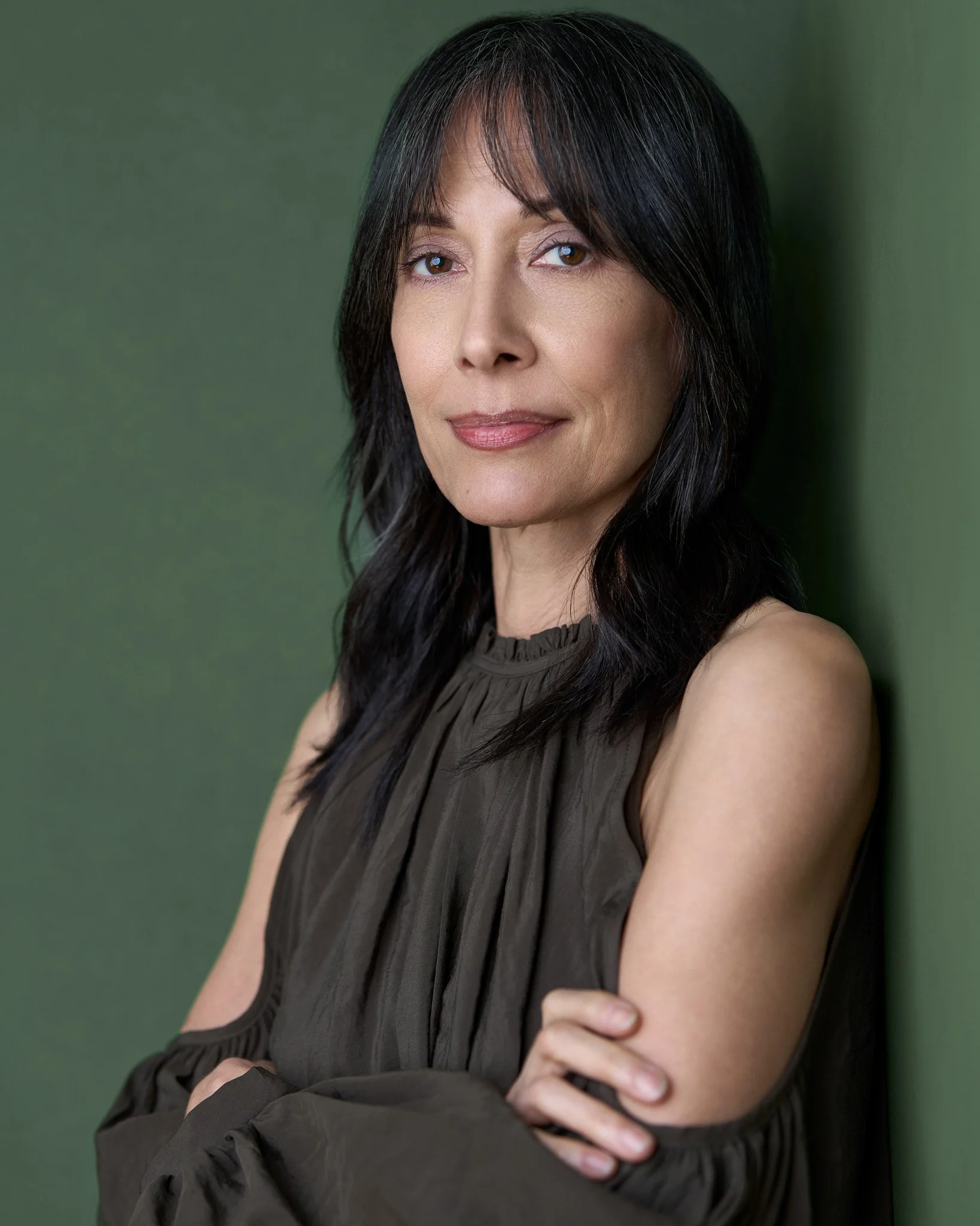 Acting headshot of a woman with long dark hair, wearing a sleeveless dark blouse, stands confidently against a green wall, arms crossed, with a calm, poised expression.