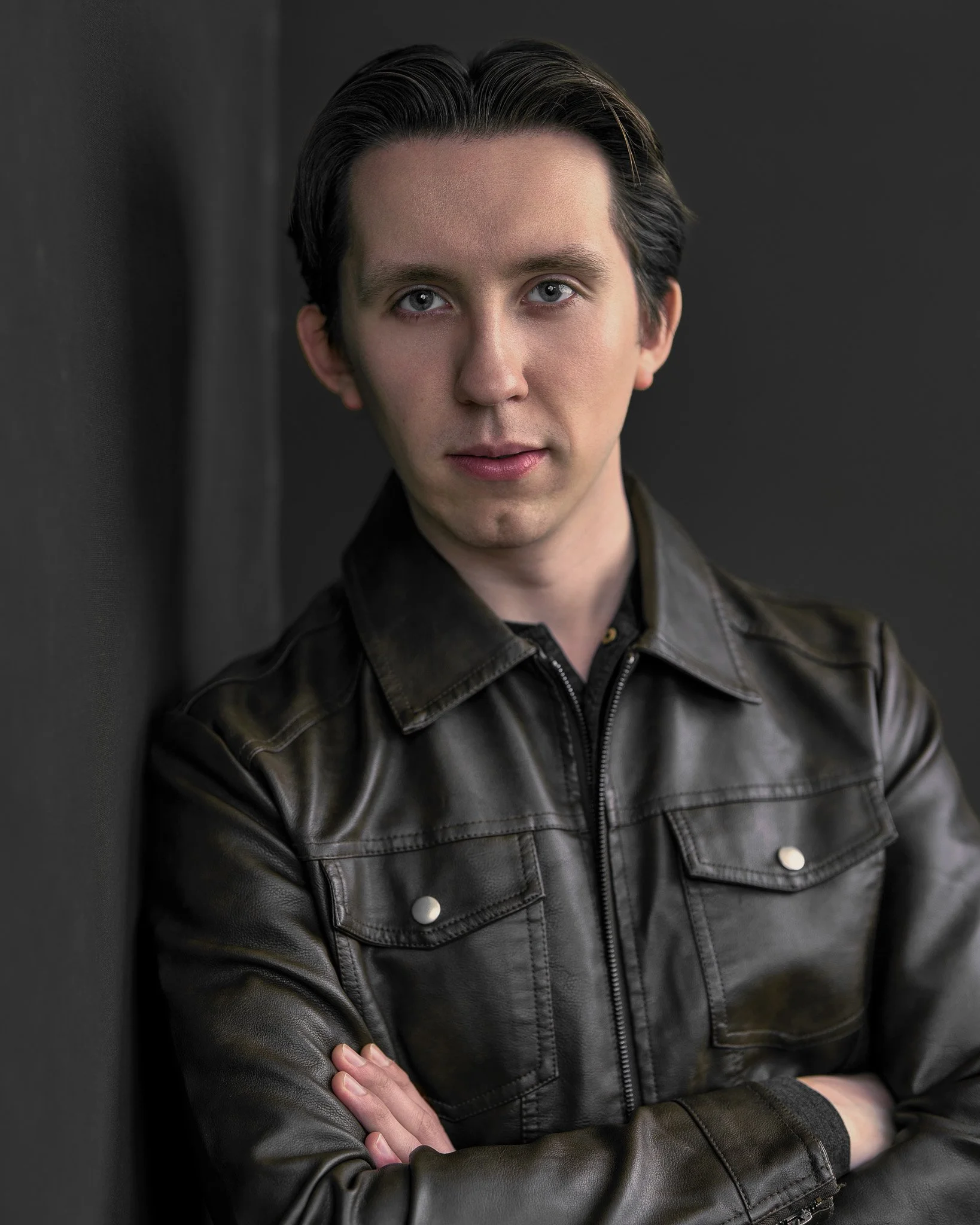 Portrait of a young man in a leather jacket stands confidently against a dark background, arms crossed. The mood is serious and introspective, with soft lighting.