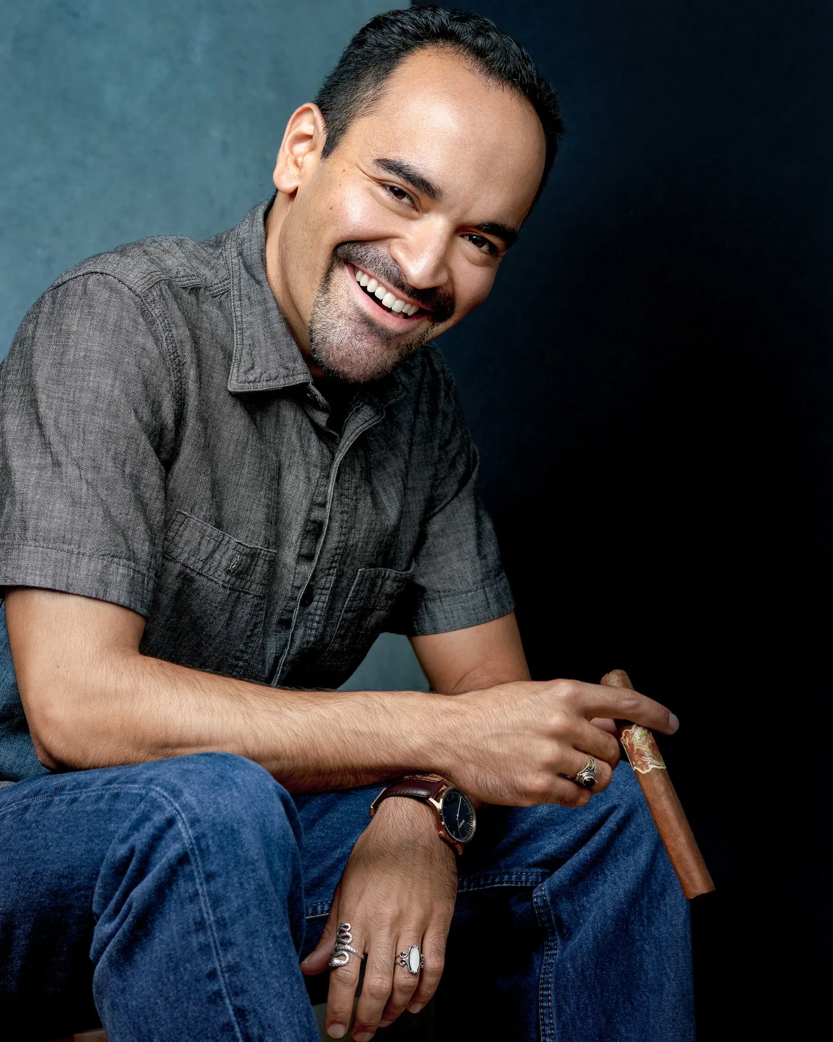 A man smiling with short dark hair, a goatee, wearing a gray shirt, jeans, a watch, and rings, sitting against a dark background.