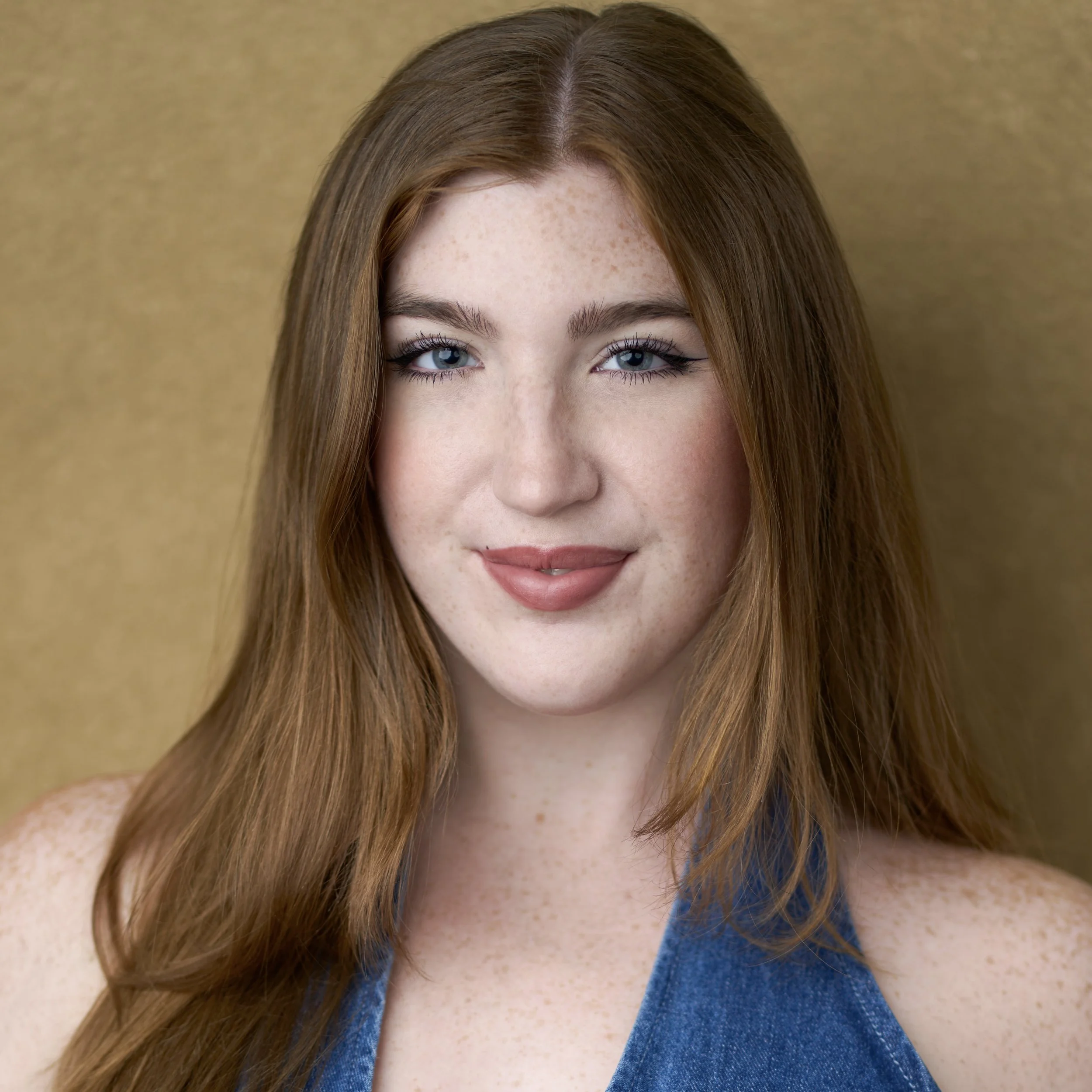 A young woman with long blonde hair and light skin, wearing a gray tank top, standing in front of a beige background.