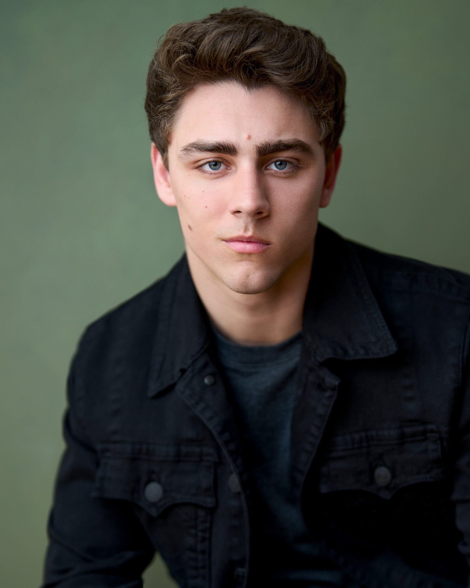 Acting headshot of a young man with short brown hair and blue eyes, wearing a black jacket over a dark shirt, gazes seriously at the camera. Green background.