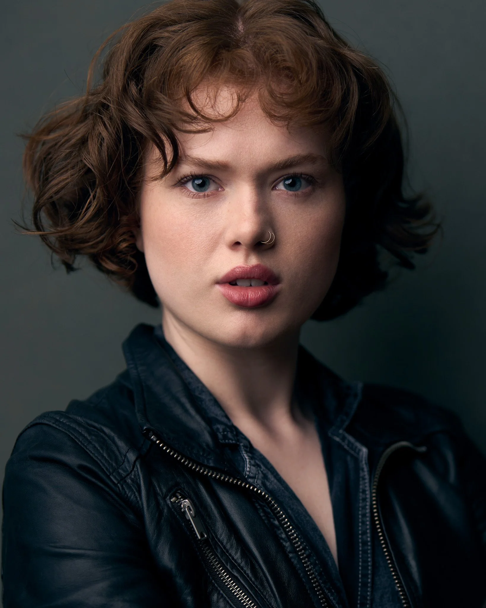 Close-up of a woman with short wavy brown hair, blue eyes, wearing a nose ring, and a black leather jacket against a dark background.