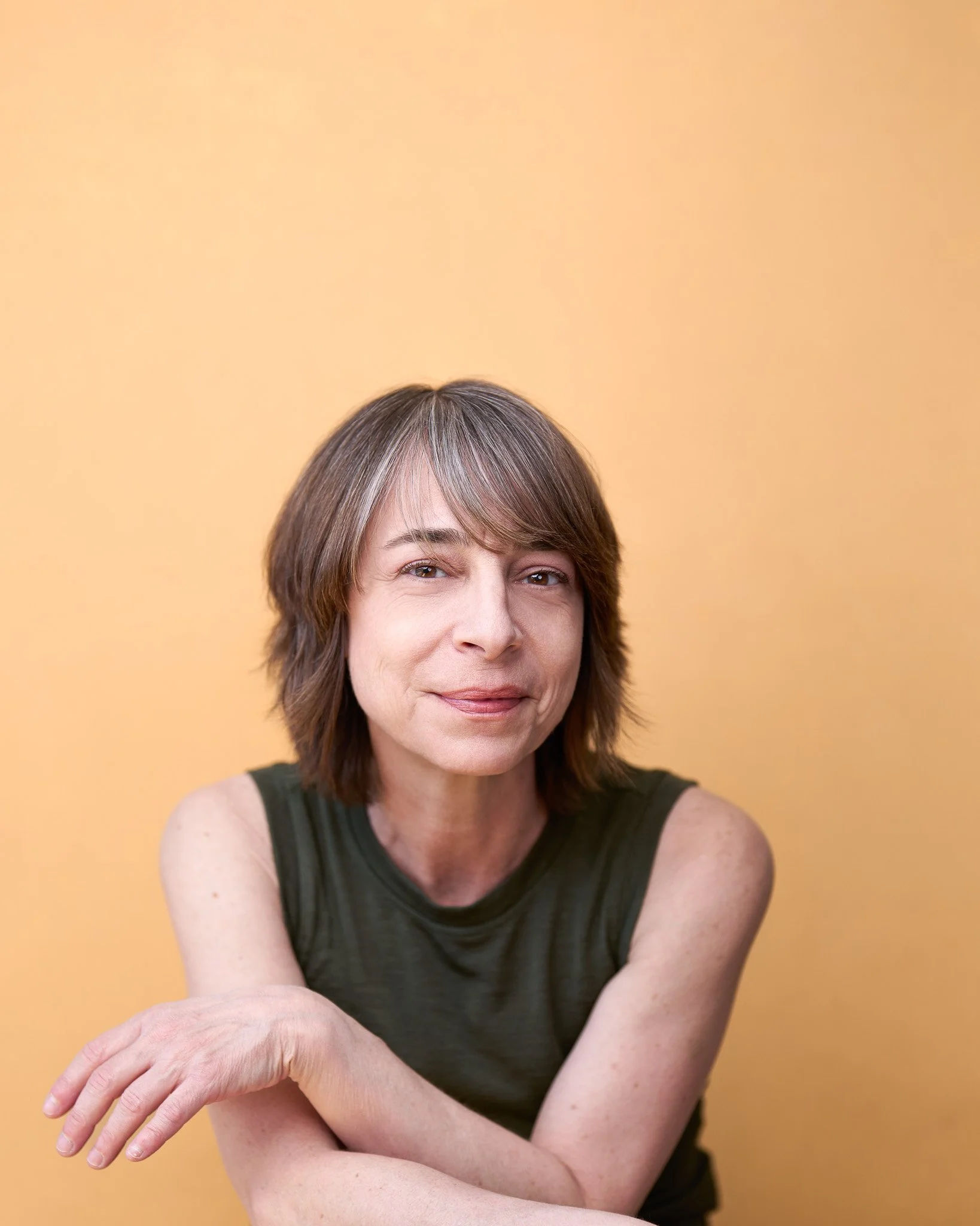 A portrait of a woman with shoulder-length brown hair, smiling slightly, wearing a sleeveless dark green top, against a solid peach background.