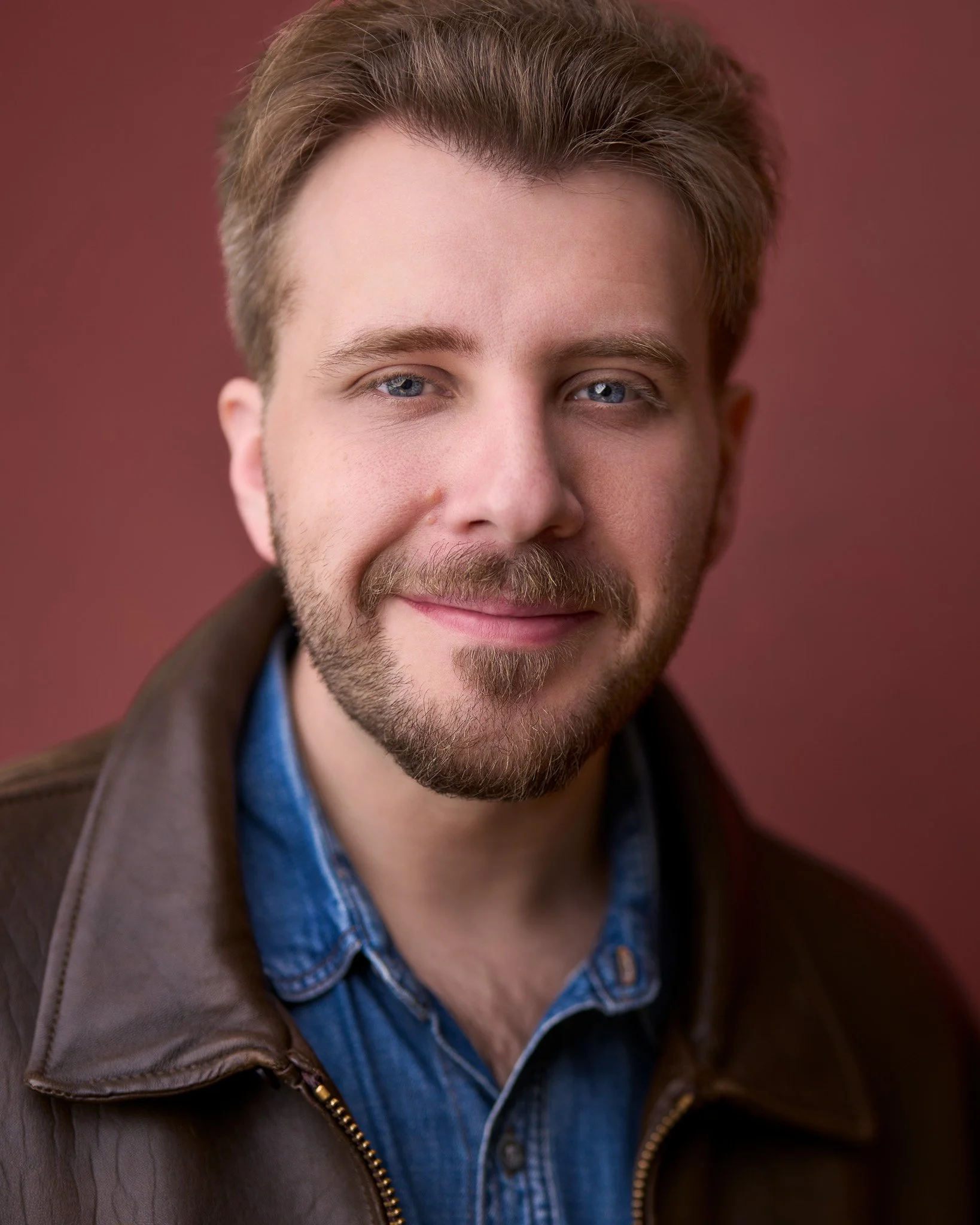 Acting headshot of a man's face with blue eyes, brown hair, and a beard, smiling slightly, with a maroon background.
