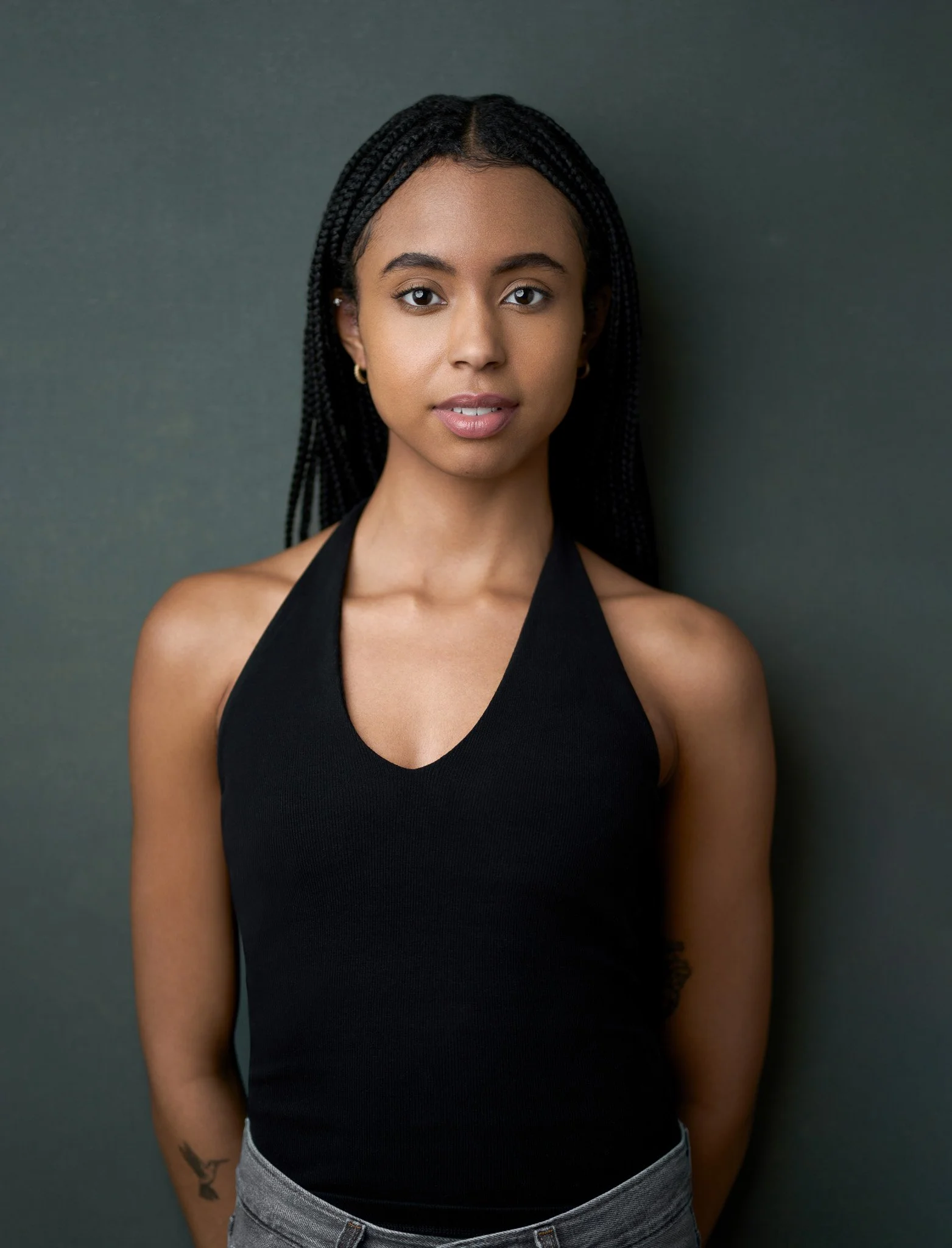 Acting headshot of a young woman with braided black hair, wearing a black sleeveless top, standing against a gray background.