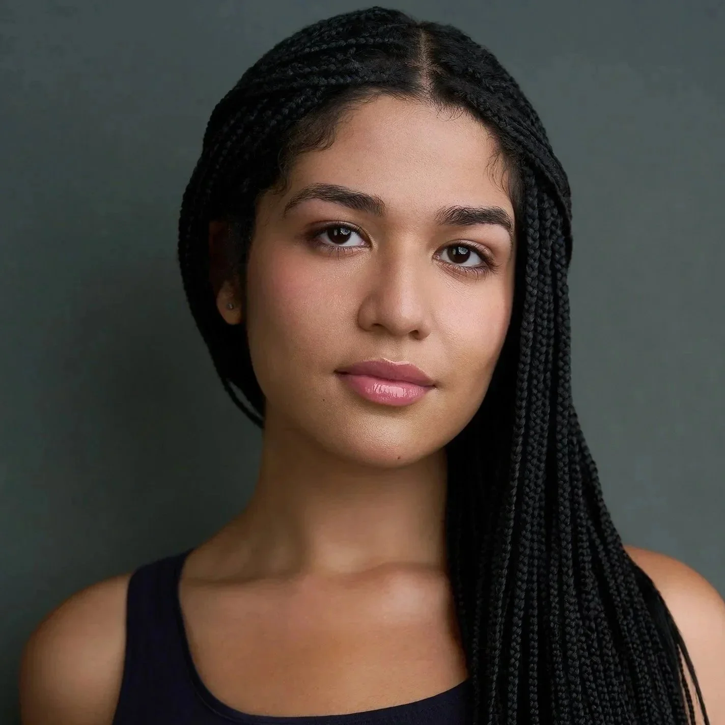 Close-up of a young woman with dark braided hair, wearing a black top with a V-neck, standing against a plain dark background.